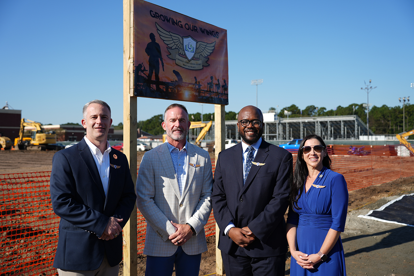 Four people in suits and dresses stand in front of a construction site sign.
