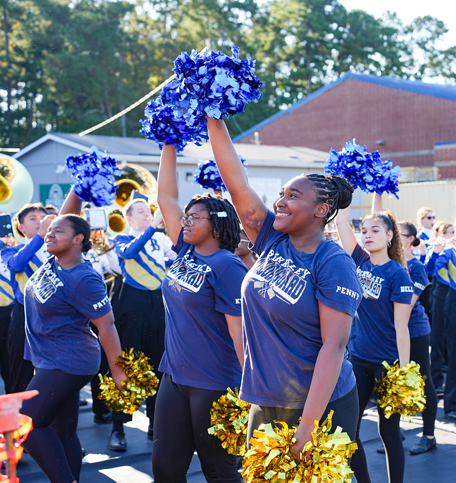 Several students in matching uniforms march outside. They hold blue and gold pom-poms. Trees and buildings in the background.