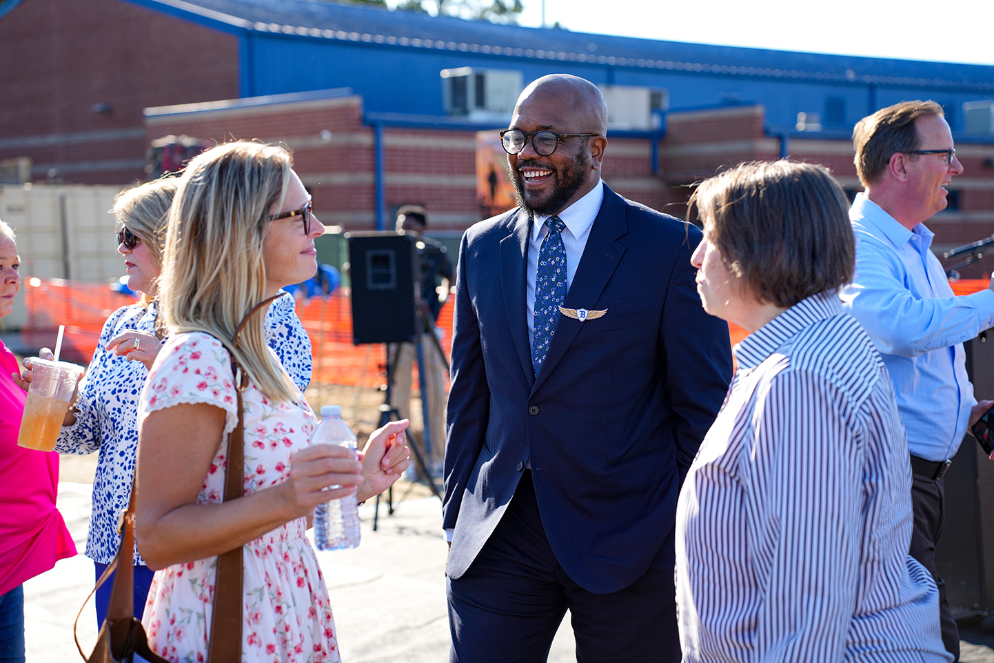 Adults in business attire conversing outside a blue building with a large window. Behind them, a tripod holds a camera.