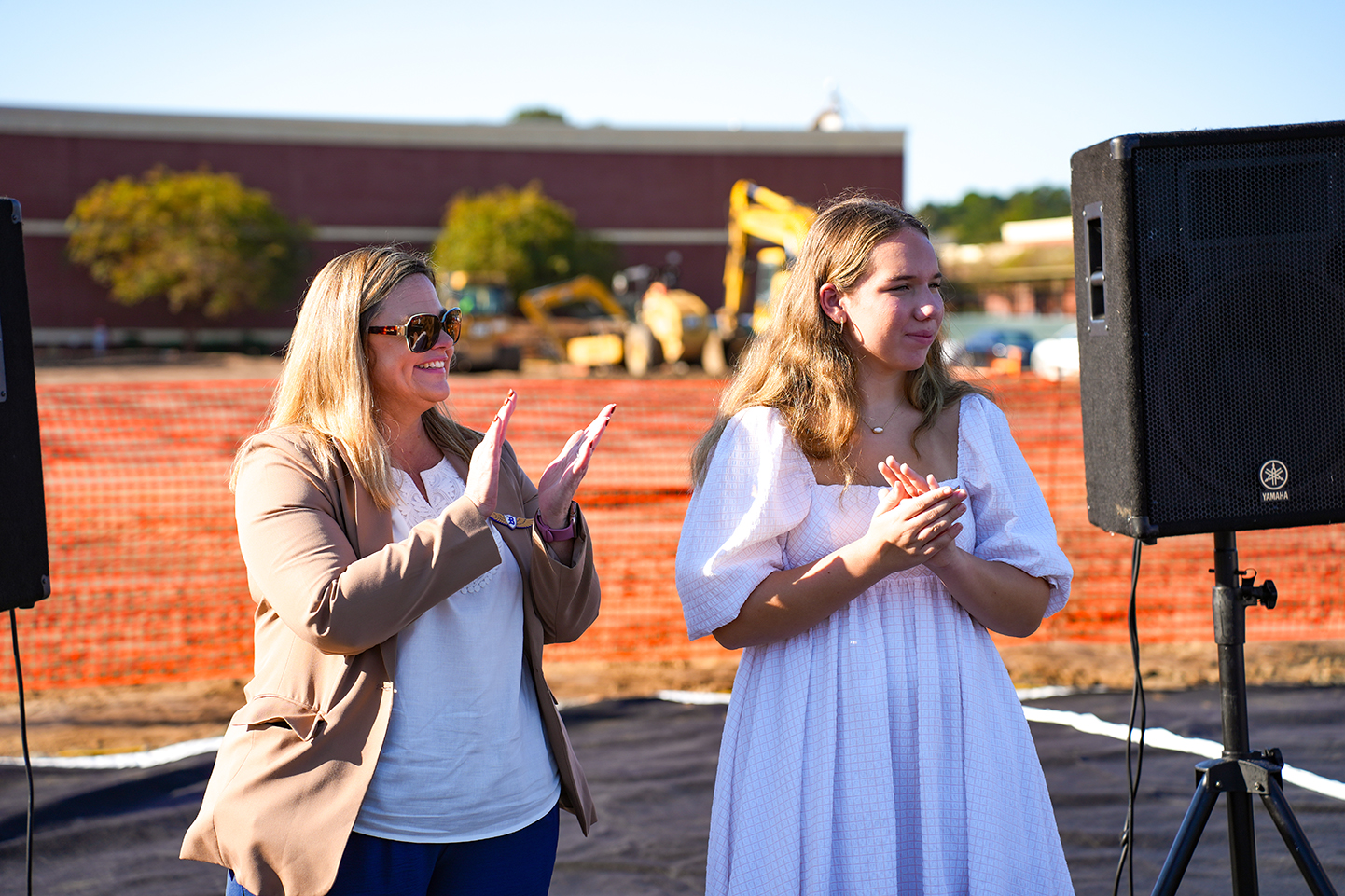 Two women clap in front of a black speaker on a tripod. Behind them, construction equipment is visible.
