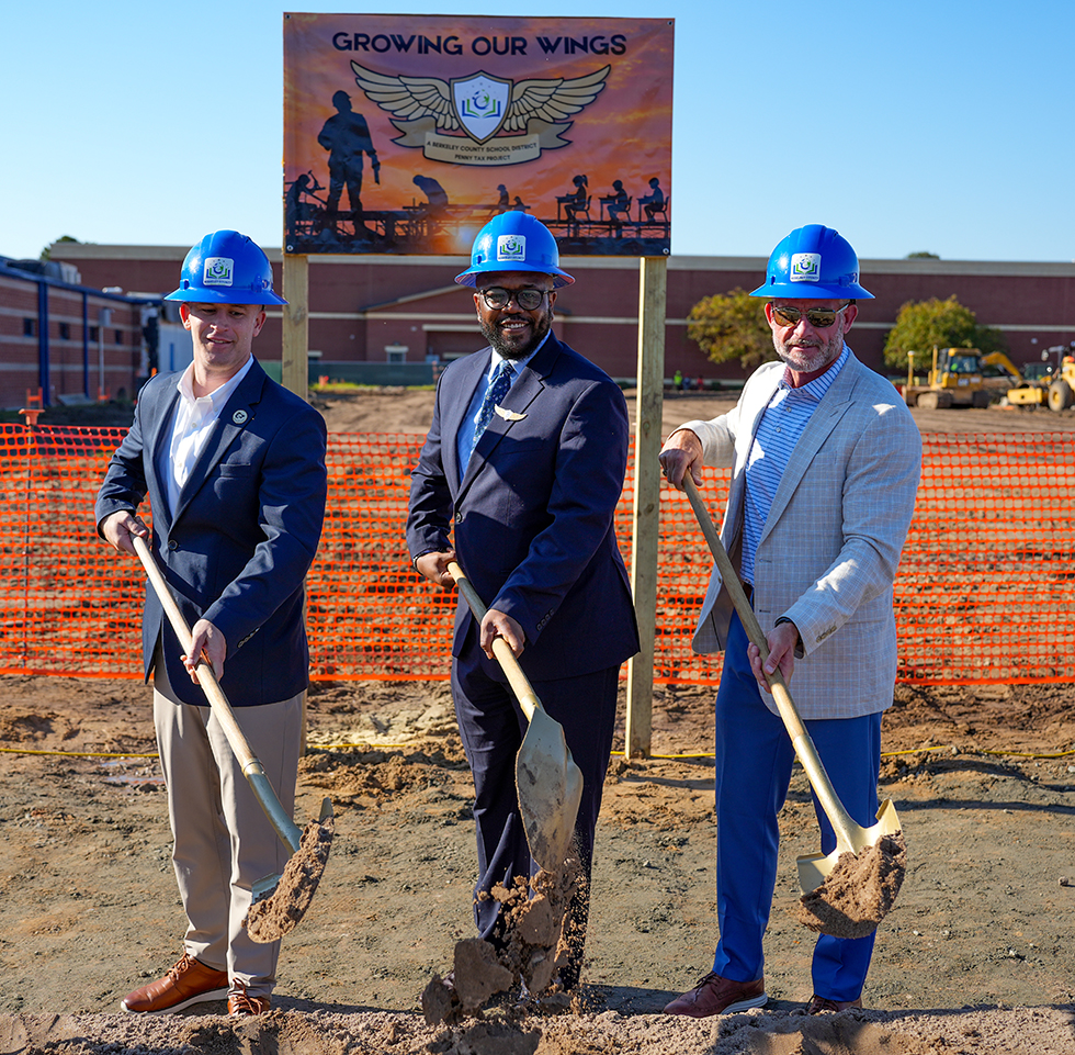 Three men in suits and helmets hold shovels at a construction site, with a banner reading "Growing Our Wings" behind them.
