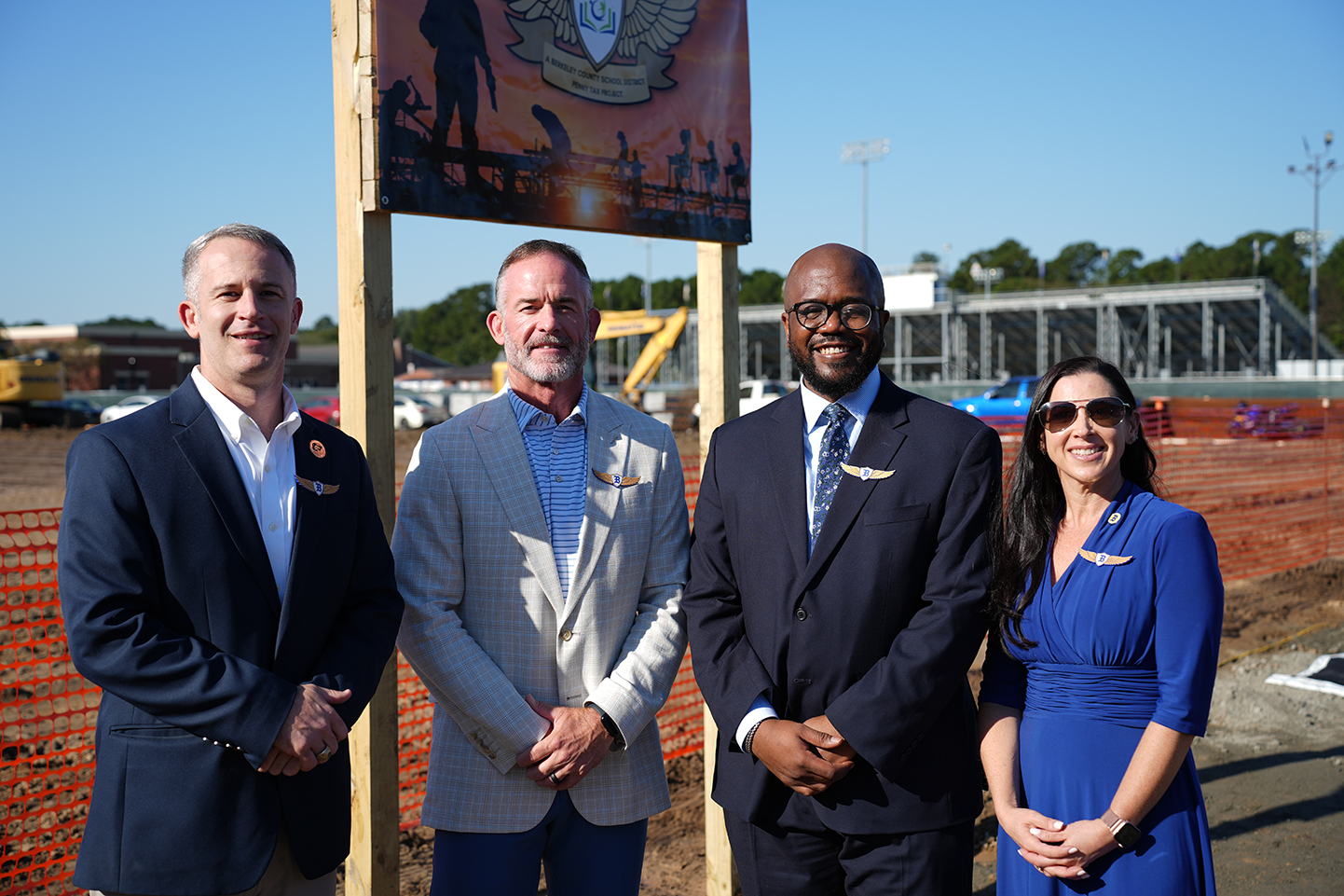 Four people standing behind a sign on a construction site with a partially built stadium in the background.