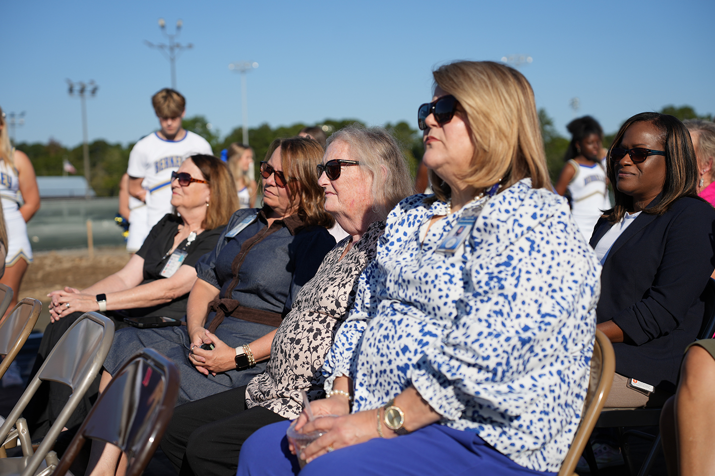 Adults sit in folding chairs watching a group of children perform on a field in a schoolyard.
