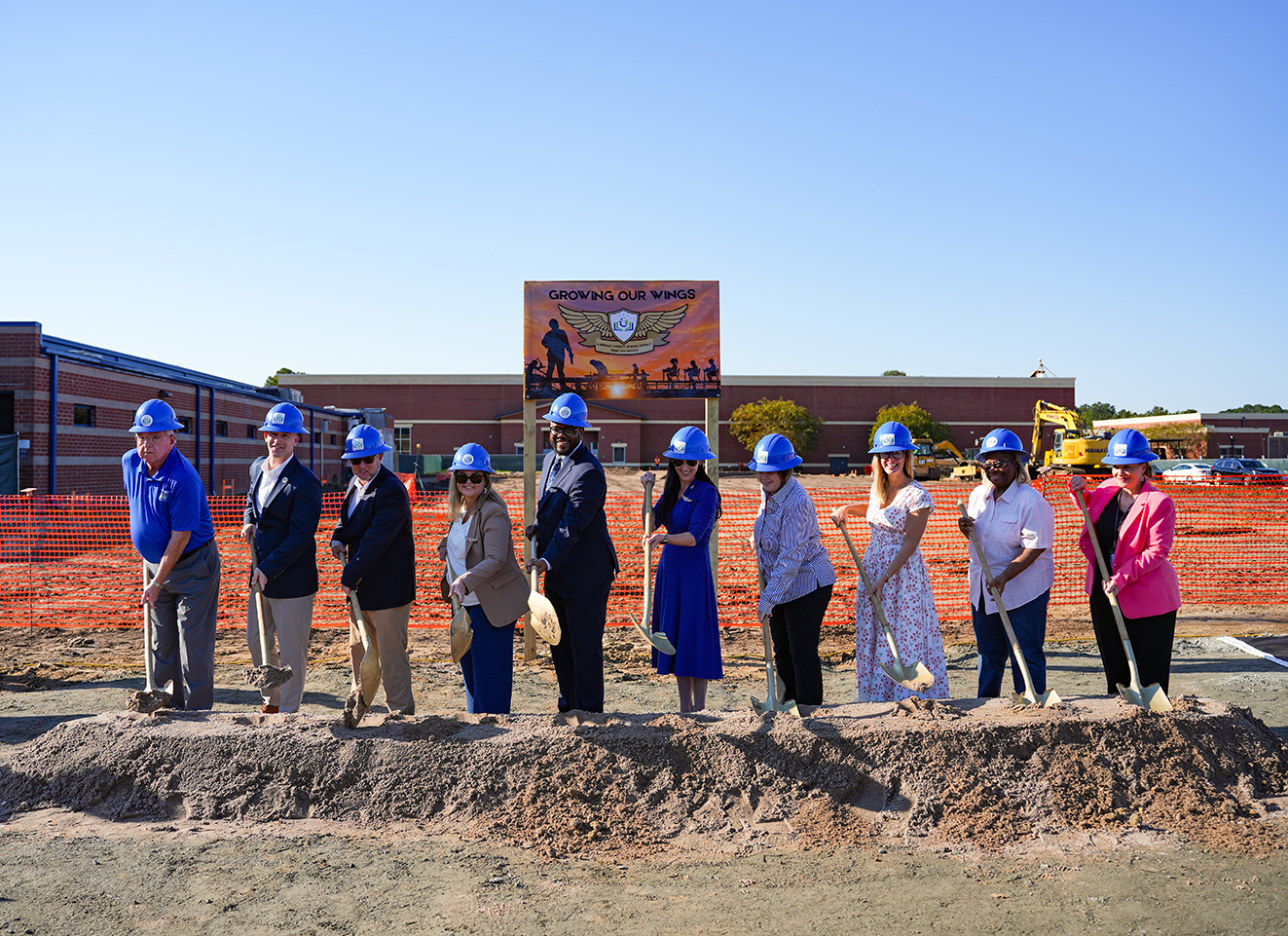 People in hard hats shovel dirt in front of a school building. Behind them is a sign and an orange fence.