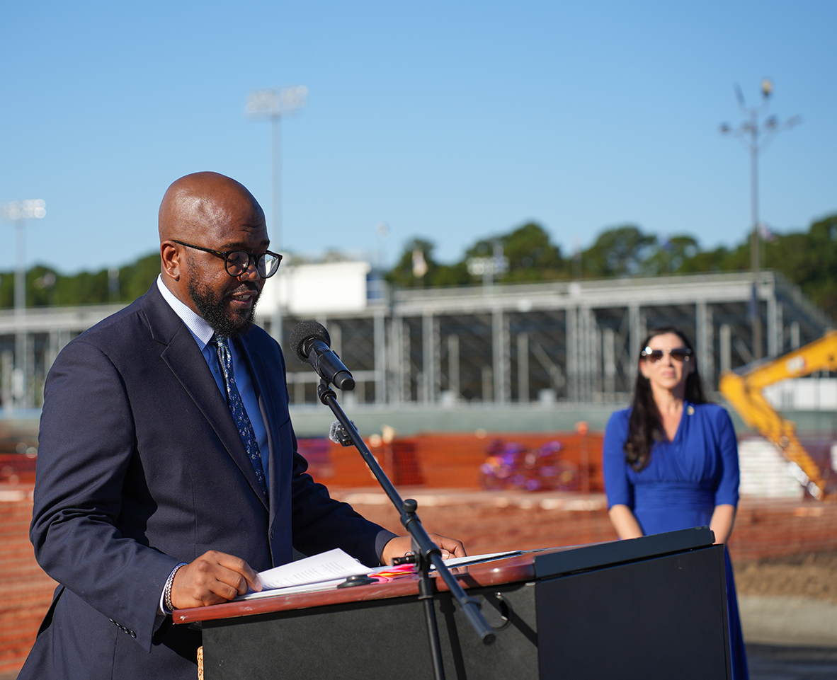 Person speaks at podium; another person stands nearby. Papers, microphone, and construction site in background.