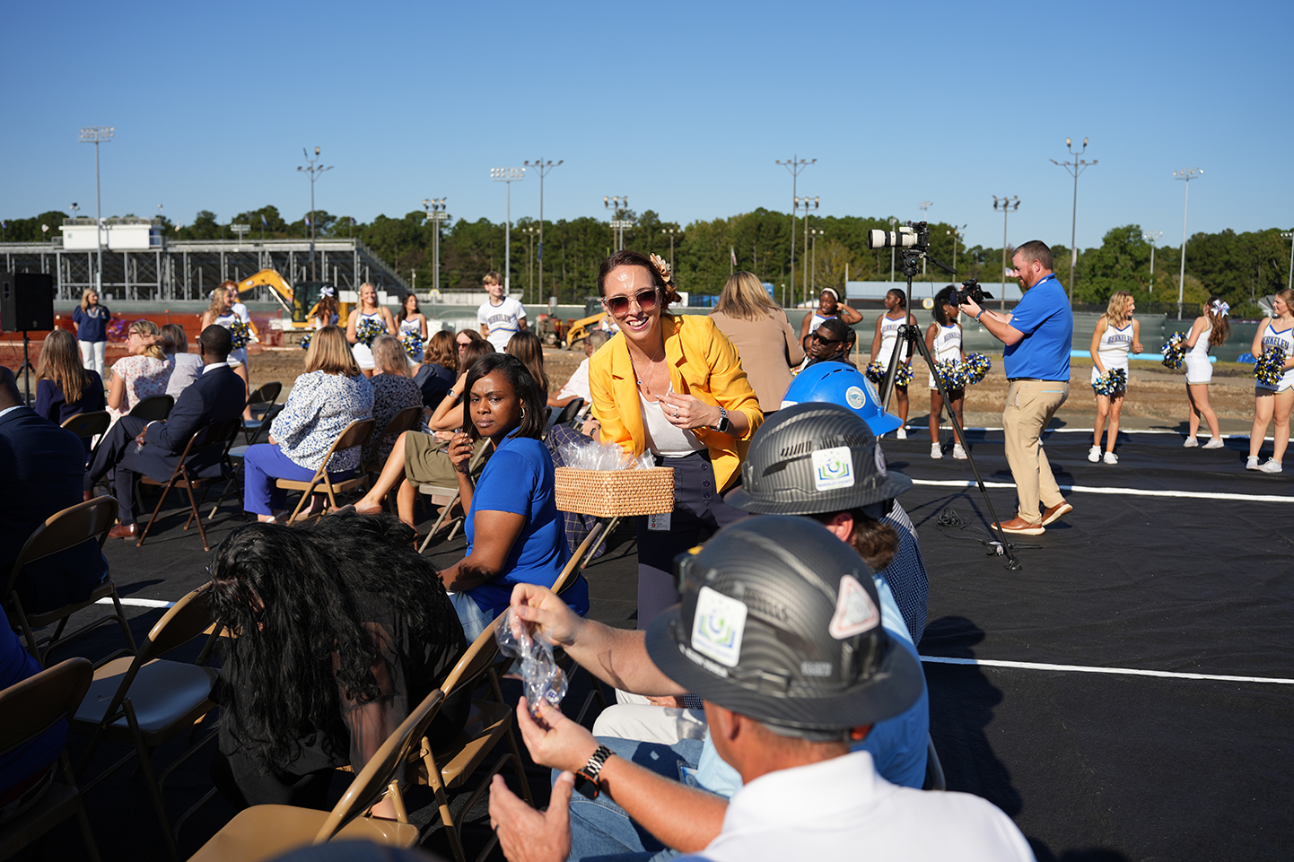 People in hard hats and some in blue shirts sit on chairs on a track. A woman holds a box.