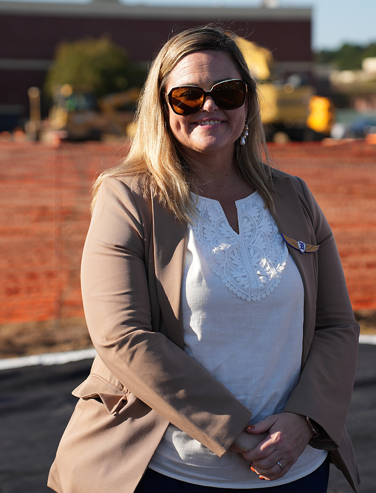 Woman wearing sunglasses, a white blouse, and a tan blazer stands in front of a construction site.