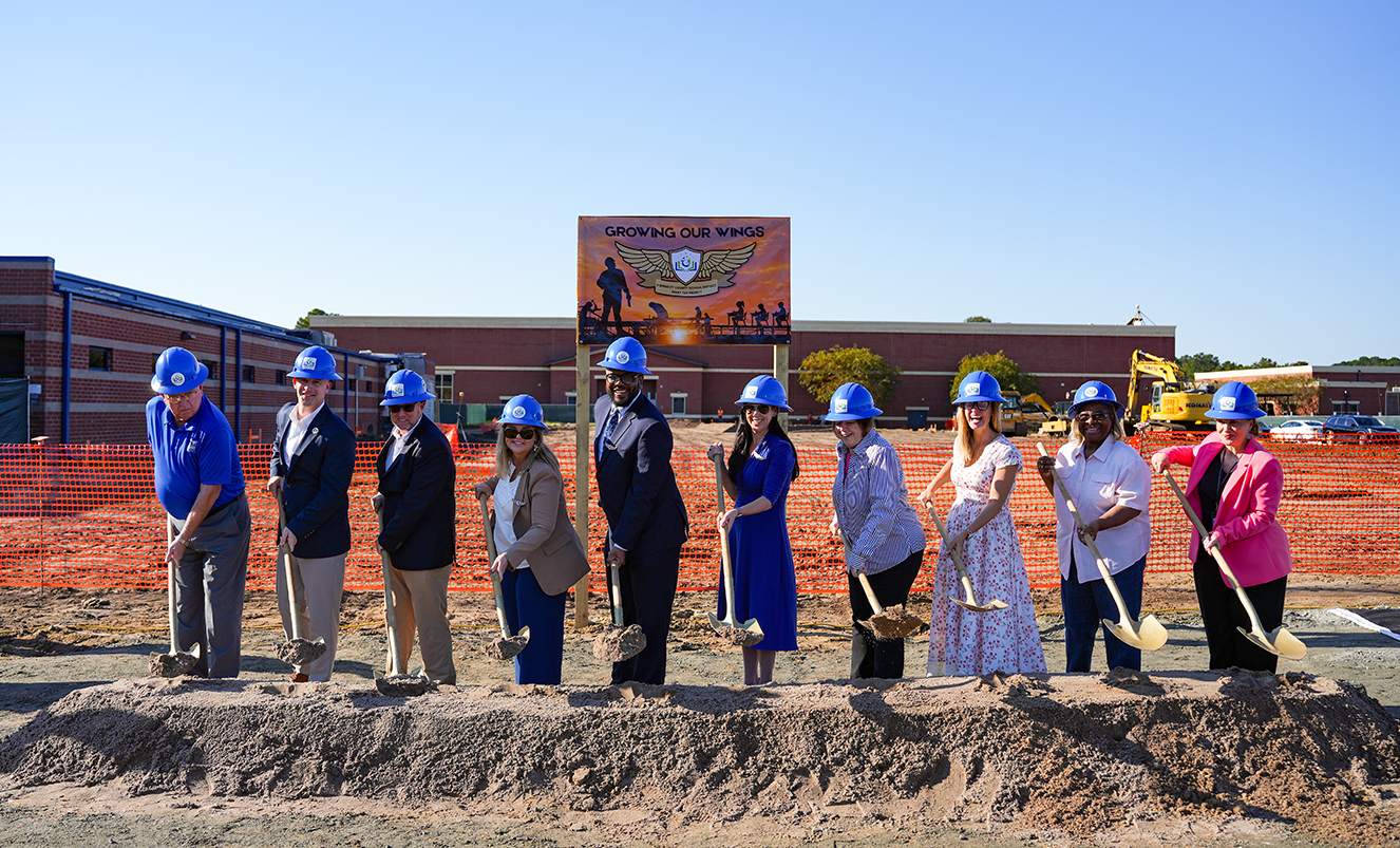 A group of people wearing hard hats shovel dirt. A building under construction is in the background.