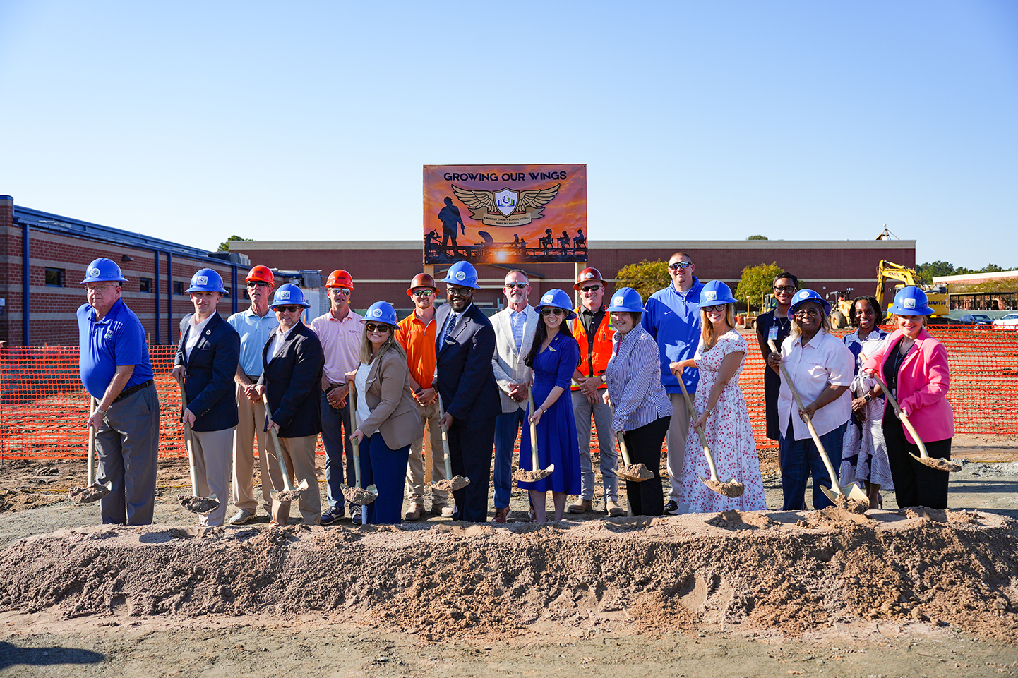 People in hard hats pose with shovels over a dirt pile, with a school building behind them.