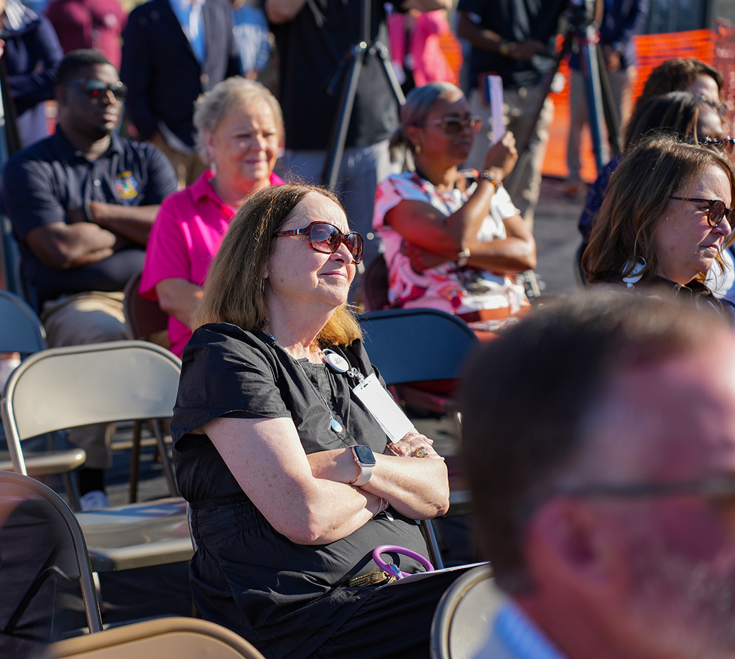 A group of adults sitting in rows of chairs, some with arms folded, others holding a badge, with some standing behind them.