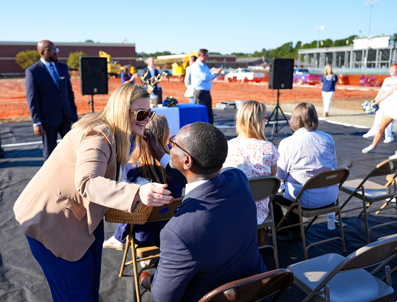 People in suits, glasses, and dresses are gathered on a field with folding chairs and a speaker.
