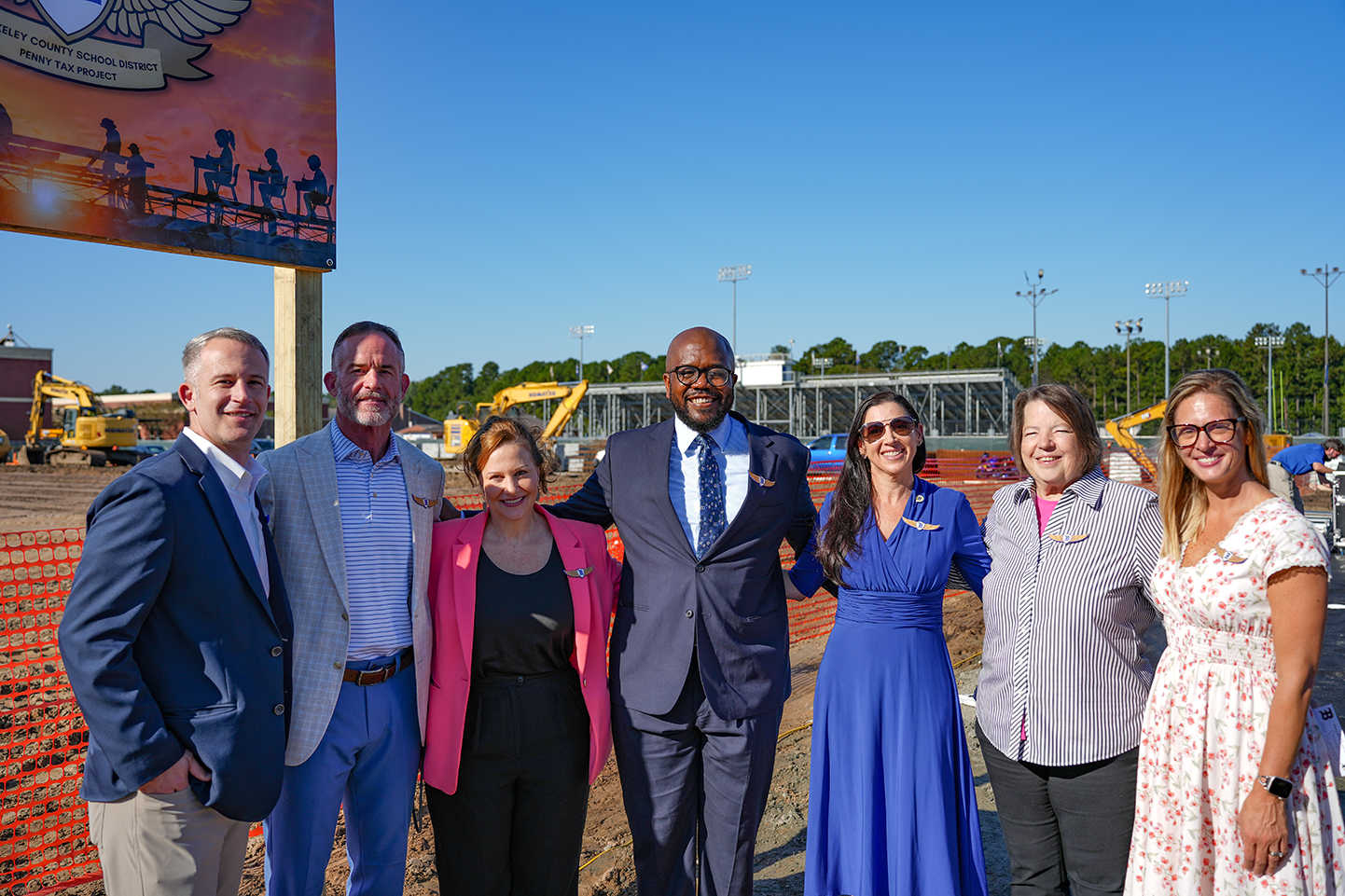 Nine adults stand outside near a large construction site with machinery, a banner, and a clear blue sky.