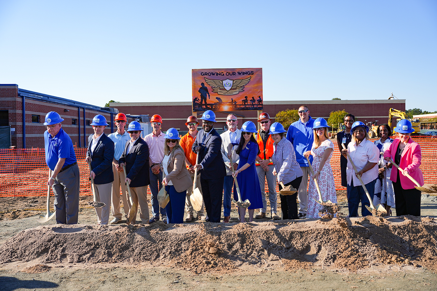 Group of people in hard hats, holding shovels, standing on a dirt pile, facing a construction sign.