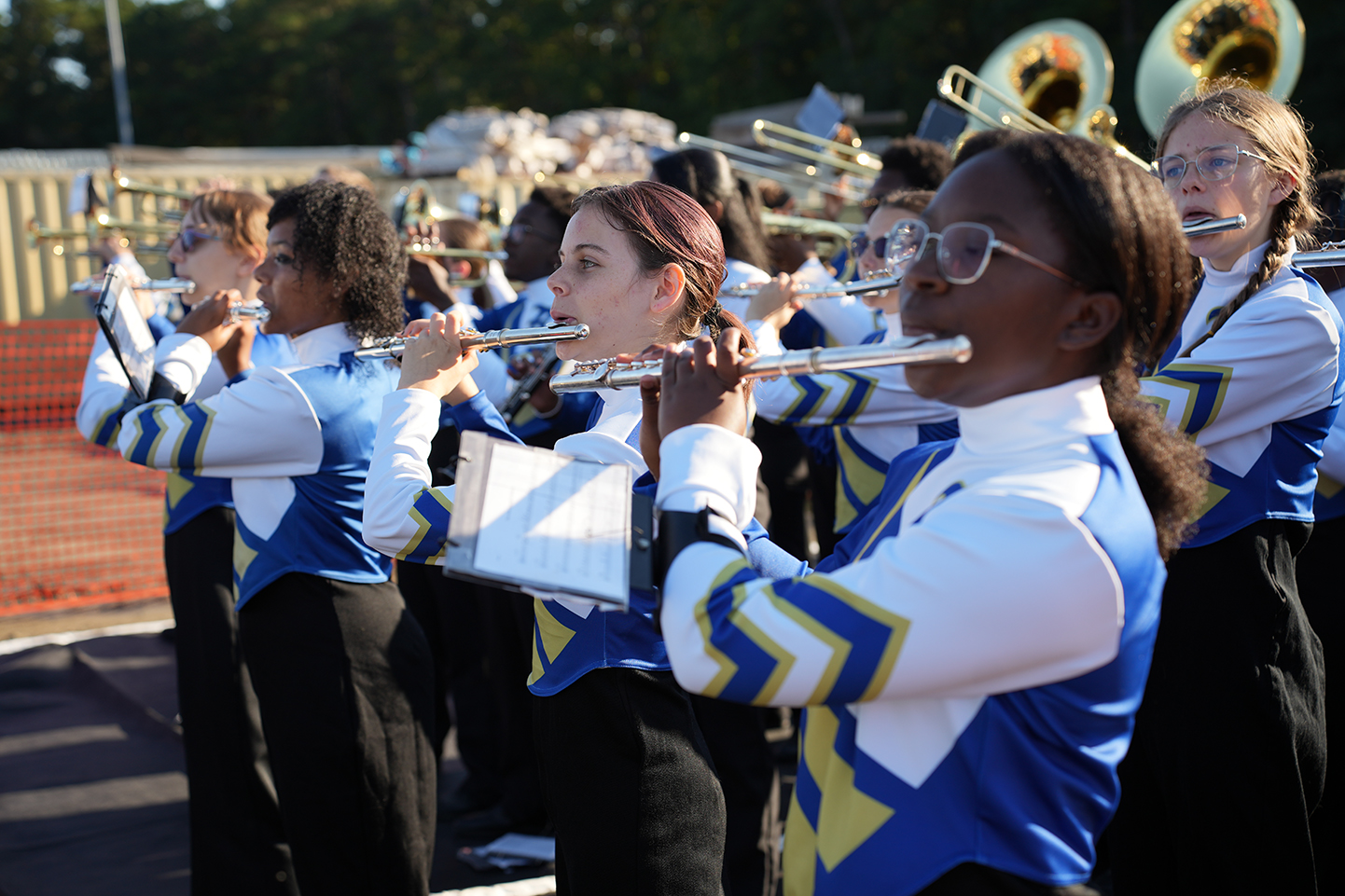 Several students in blue and white uniforms play instruments, including flutes, outdoors in front of a building.