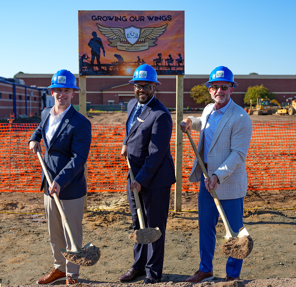 Three men with shovels stand in dirt in front of a sign that reads Growing Our Wings.