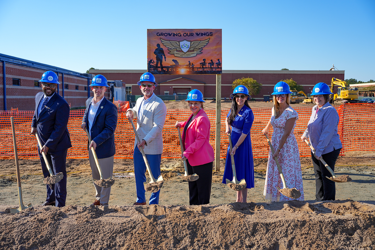 Seven people in hard hats stand in a dirt patch with shovels in hand. A sign behind them reads Growing Our Wings.
