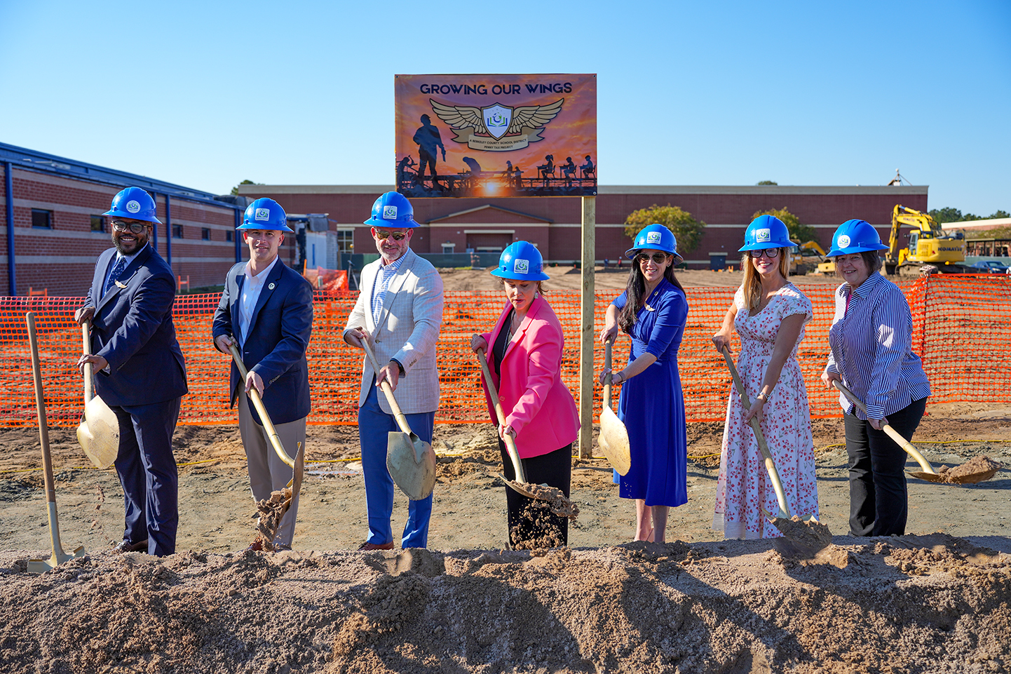 A group of people wearing hard hats hold shovels, standing on dirt mounds for a groundbreaking ceremony.