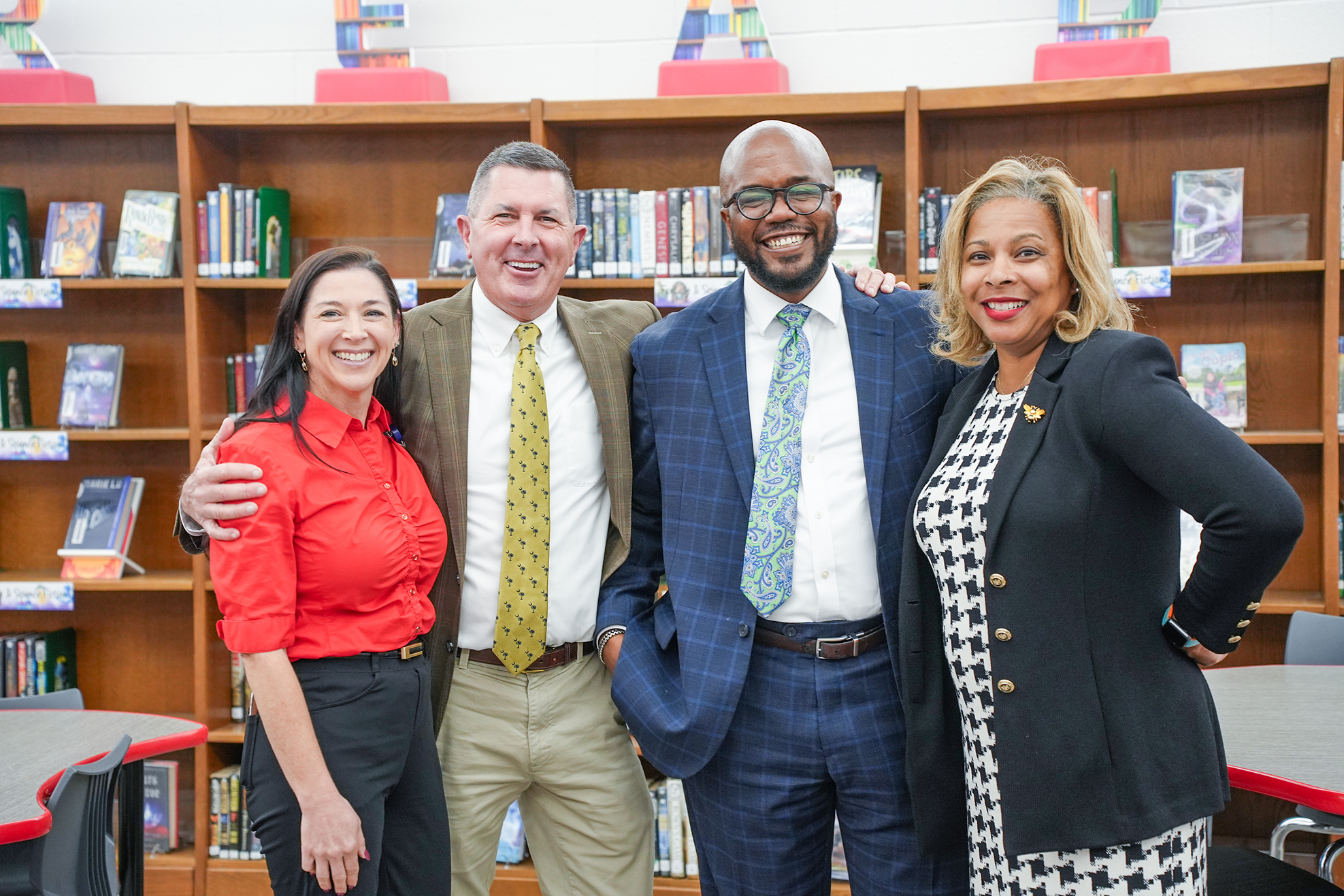 Four adults stand together in front of a bookshelf. Two are wearing red and the others are wearing blue.