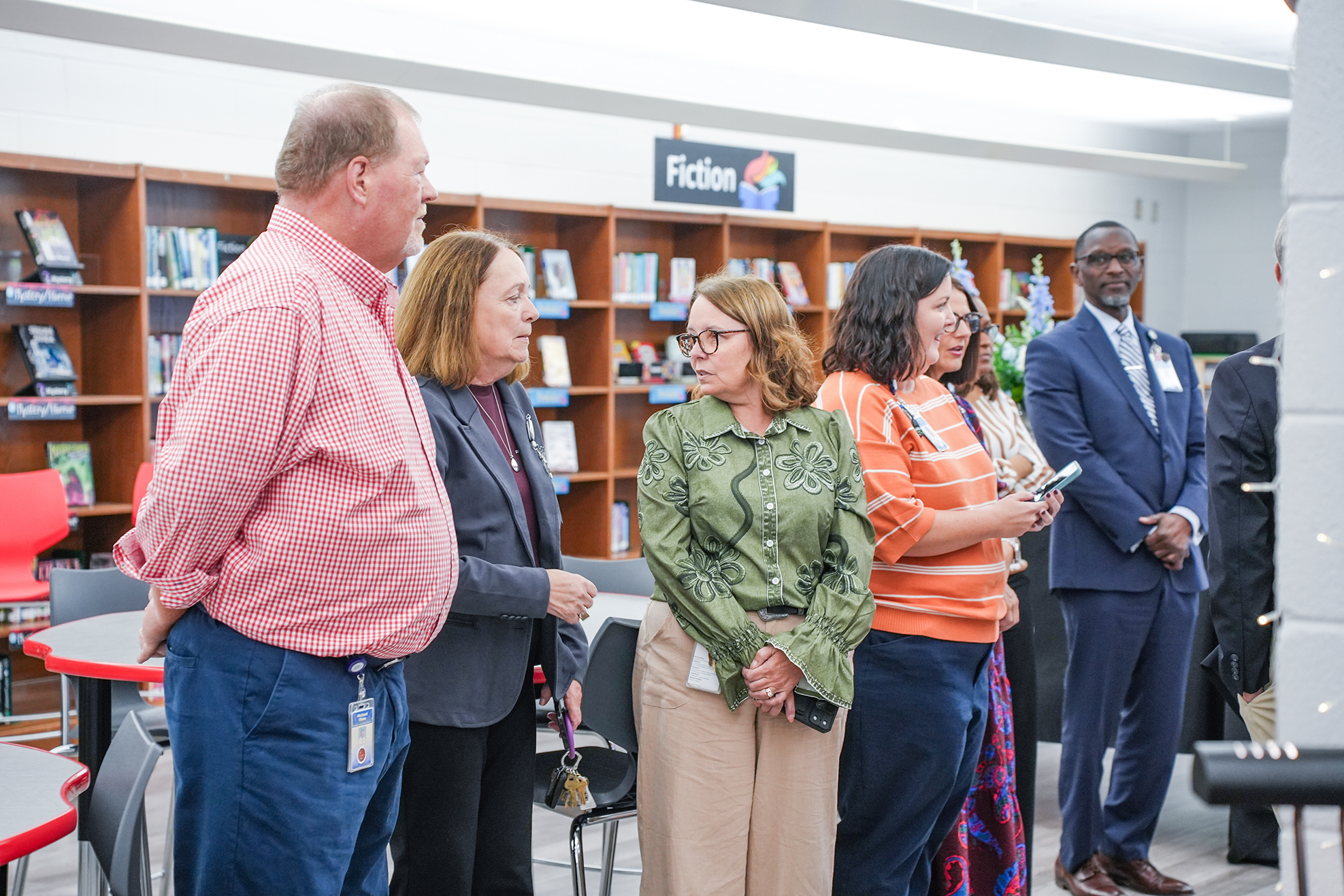 People gather in a library, standing and facing each other. Bookshelves and chairs are in the background.