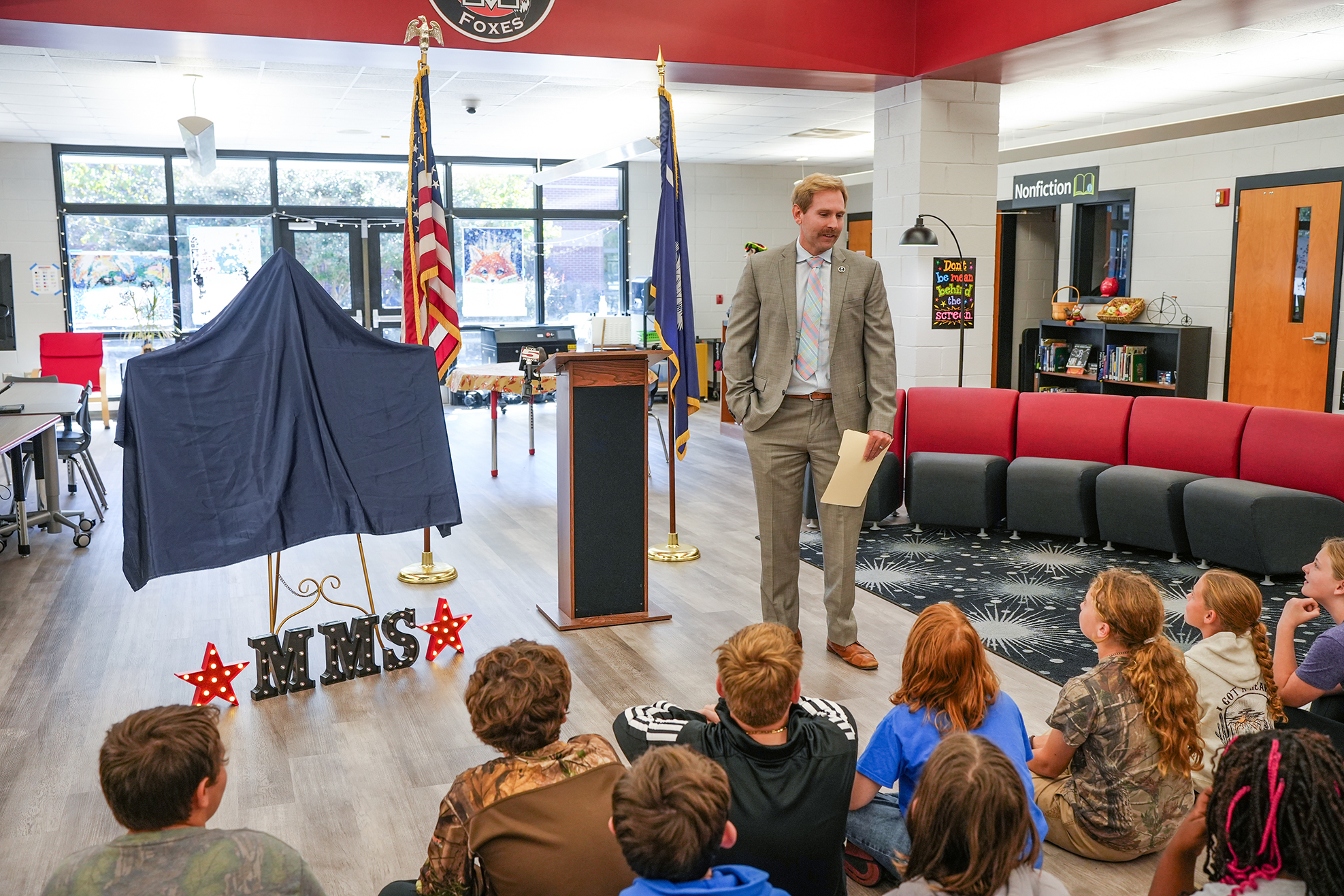 Man speaks to students seated on floor; some sit on red bench, some on black floor cushions.