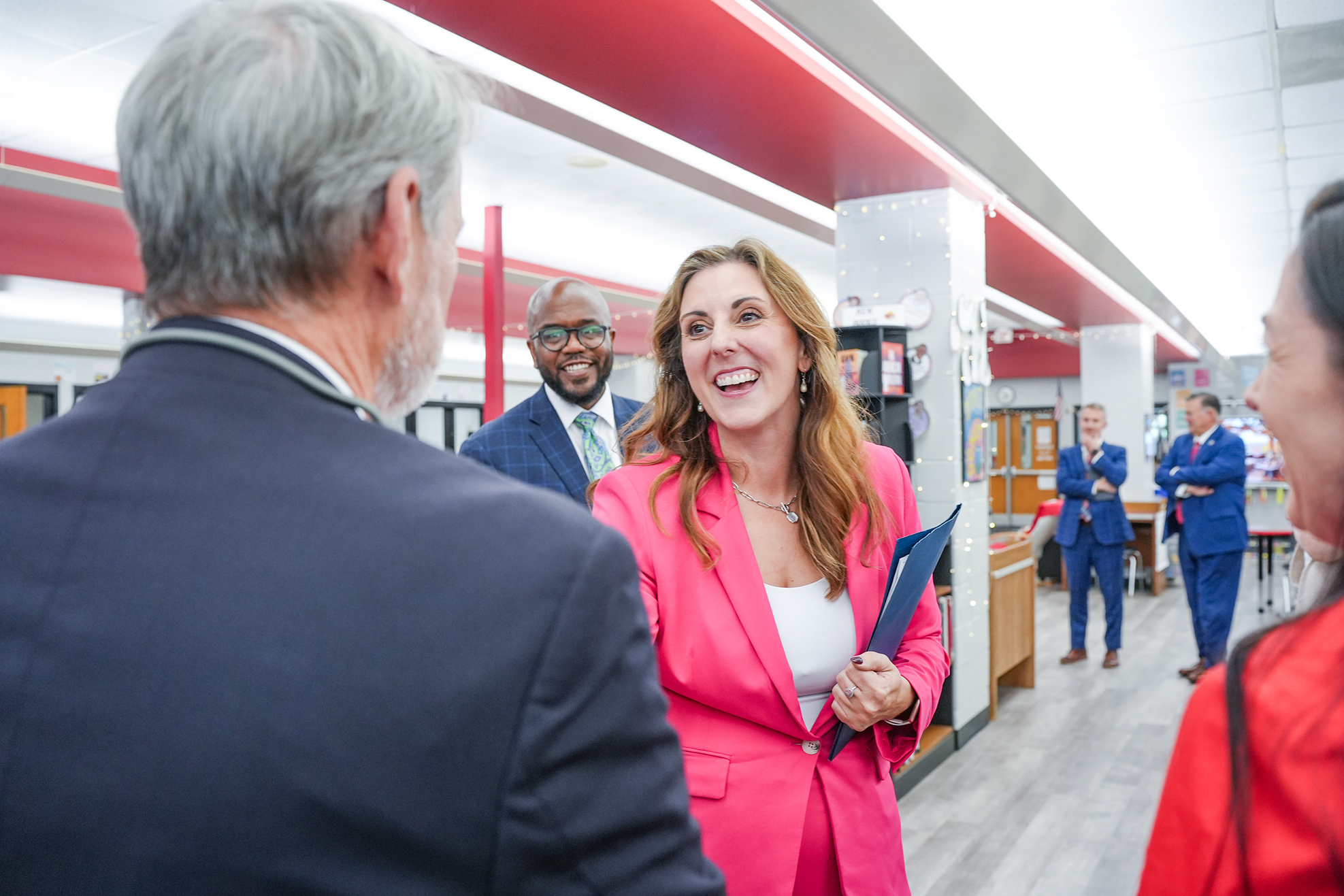 Several adults in suits and jackets talking inside a school hallway; one woman holds a folder and laughs.