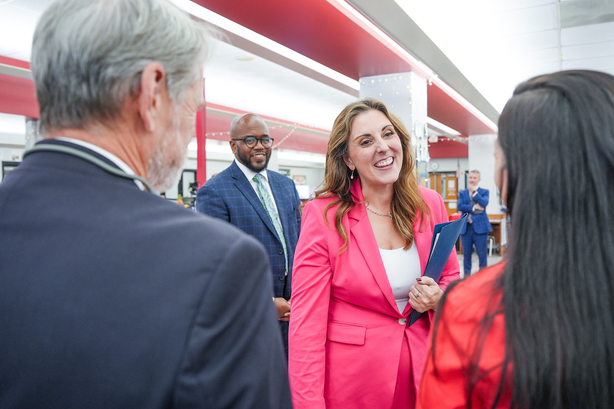 A woman in a pink blazer smiles while talking to three people in a well-lit room.