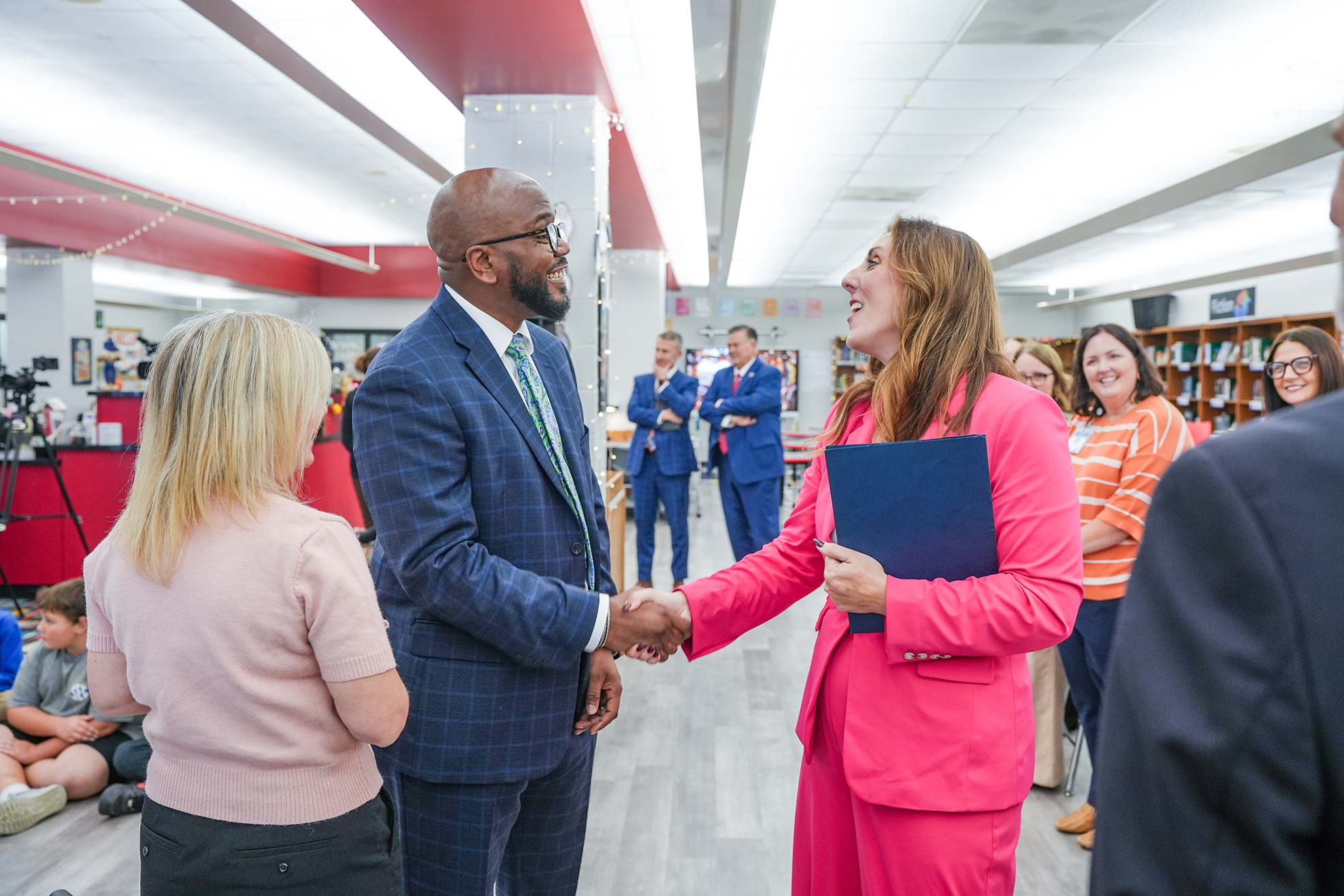 Two people shake hands in a hallway; a man in a suit and a woman in a pink suit.