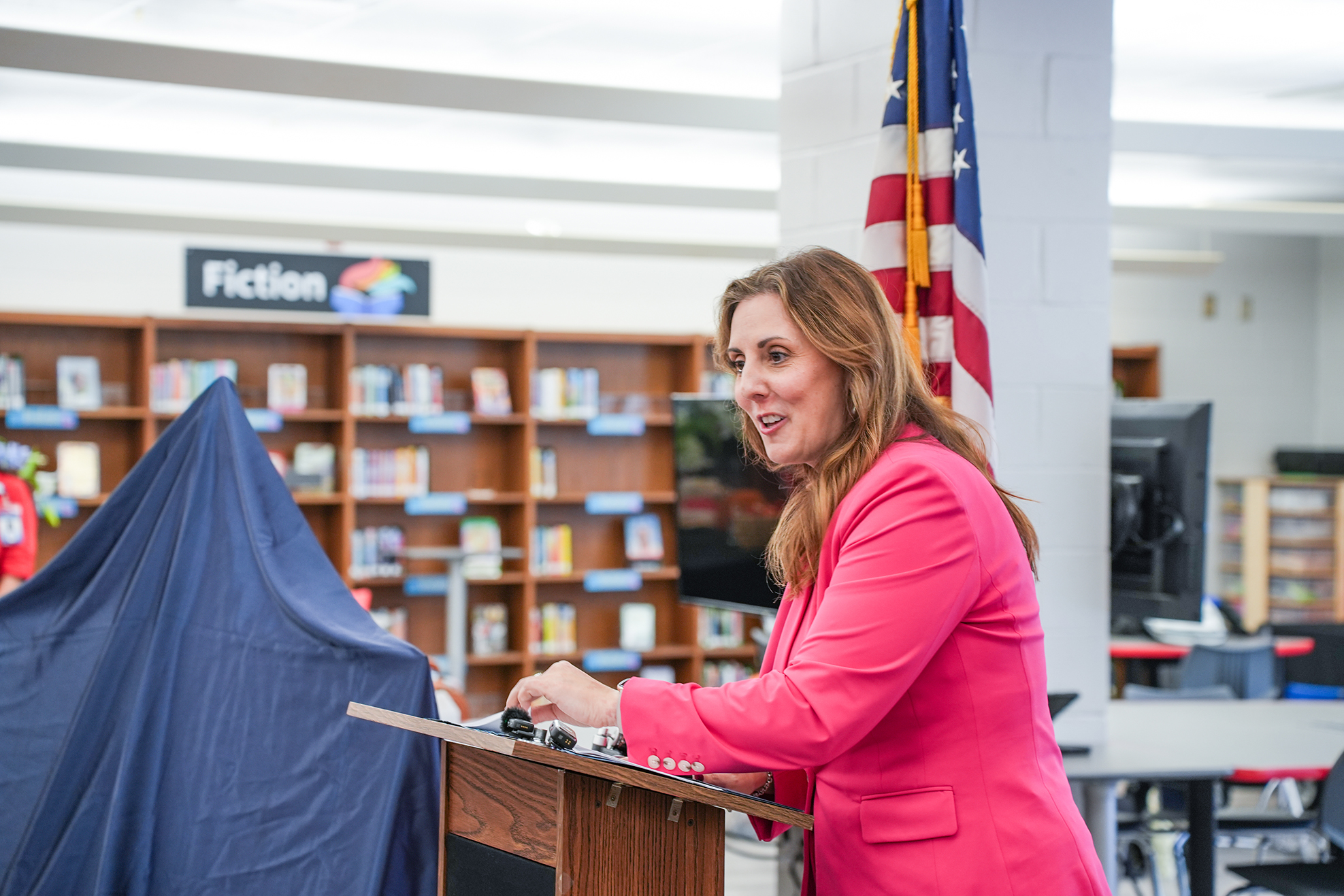 Woman in pink blazer speaking at podium in a library; blue fabric near podium; bookcases behind her.