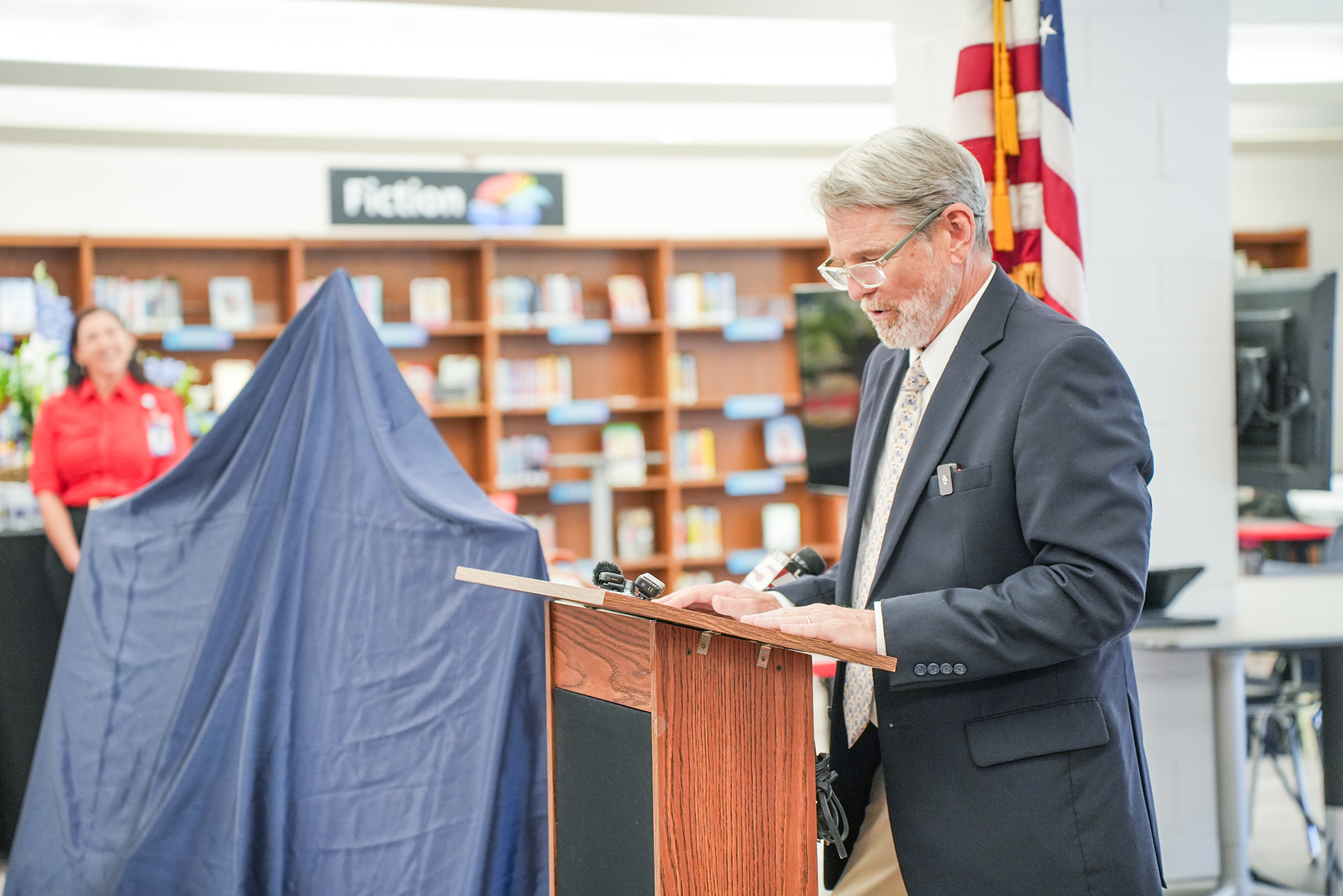 Man wearing glasses and a suit stands at podium in library, speaking into a microphone.
