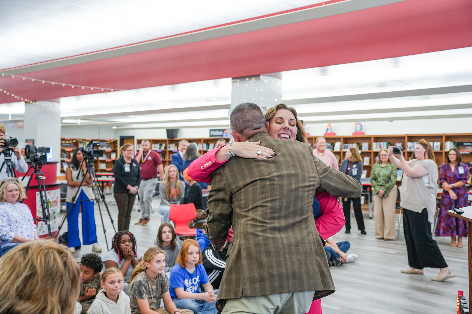 A man and woman embrace in a school library. People sit on the floor and stand around them. Some people hold cameras.