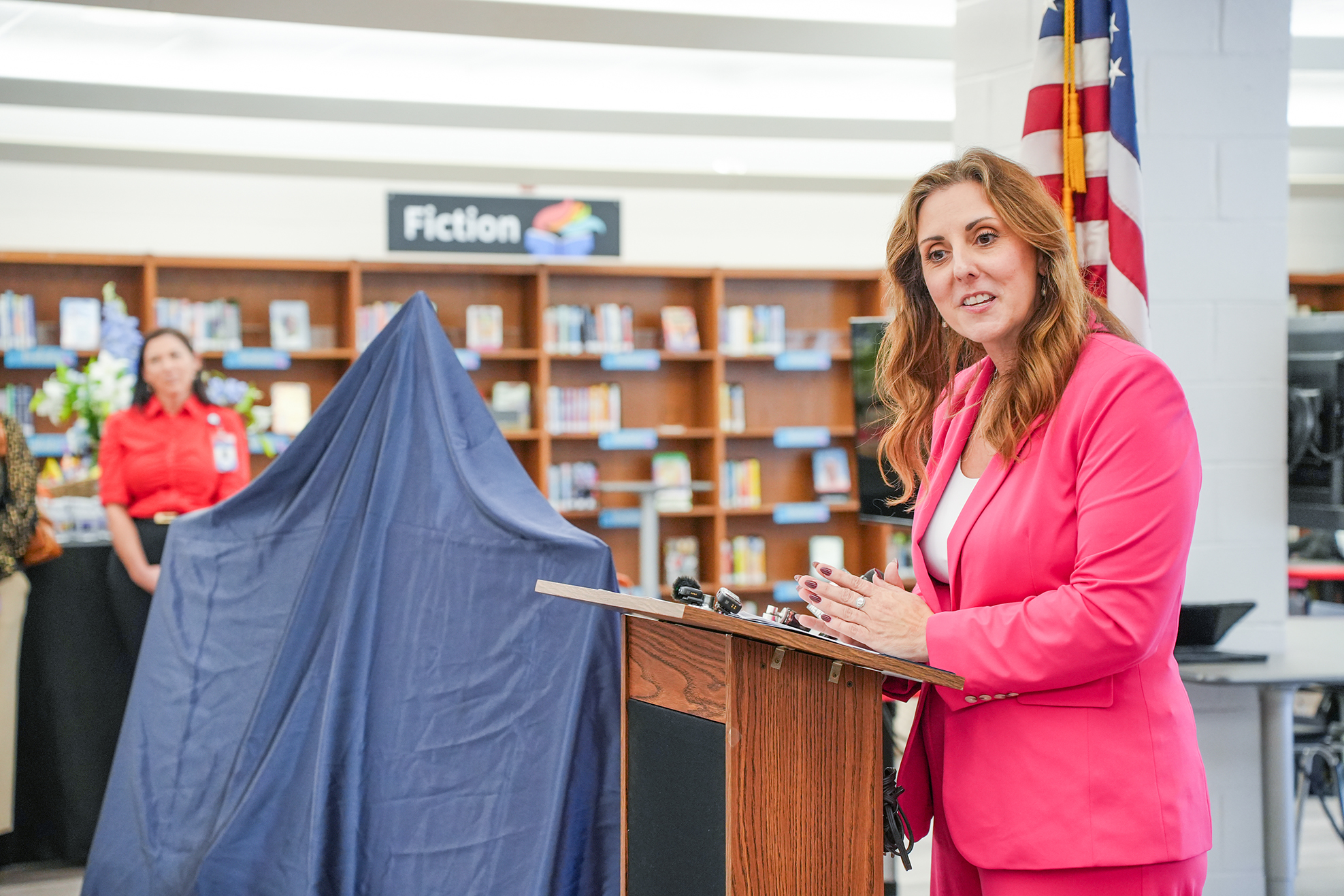 Woman at podium in library. A blue sheet covers a table behind her. Books on shelves in background.