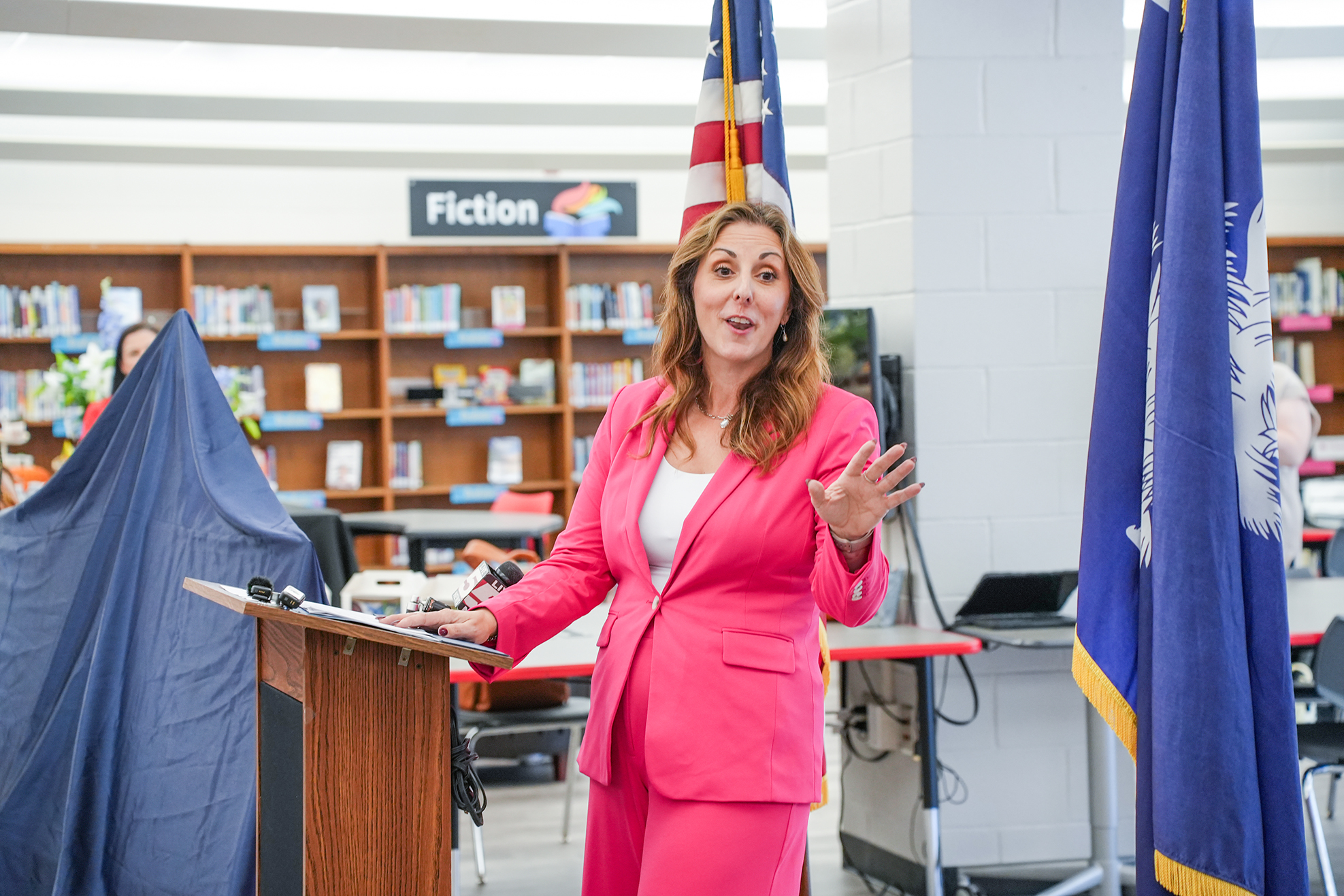 Woman speaking at podium, gesturing with hands, in front of library shelves, flag, and laptop.
