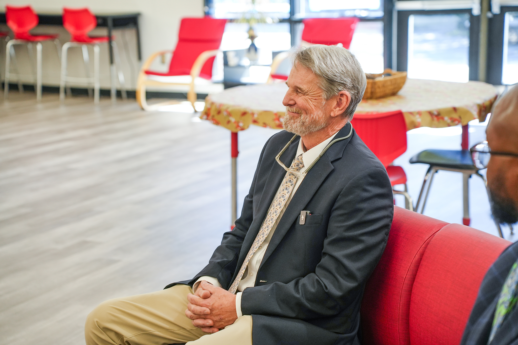 A man in a suit sits on a red sofa with folded hands, conversing with another man.