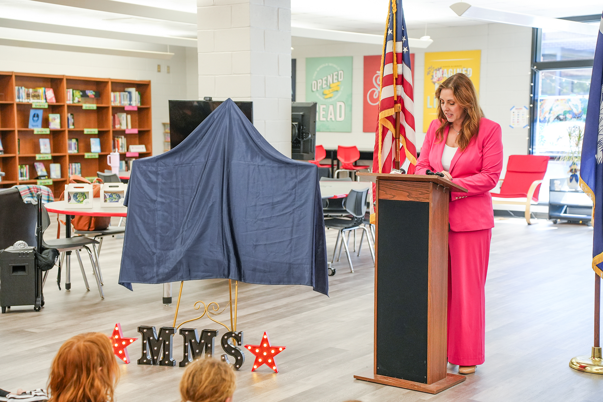A woman speaks at a podium; a blue cover is nearby, and several children listen. Behind her, bookshelves and chairs are visible.