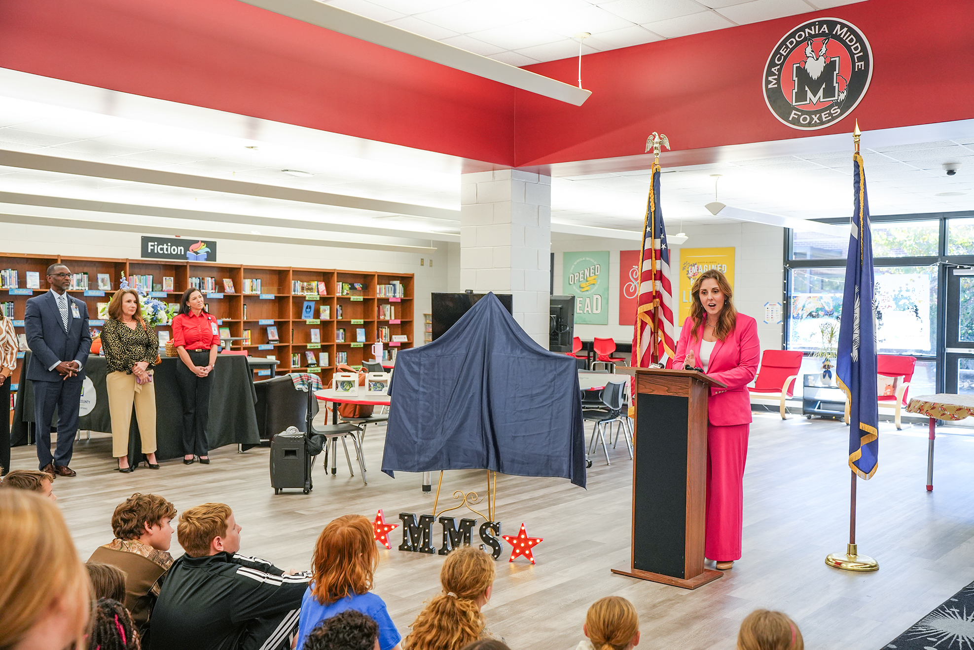 People gather in a school library. A woman stands at a podium, reading from a document.