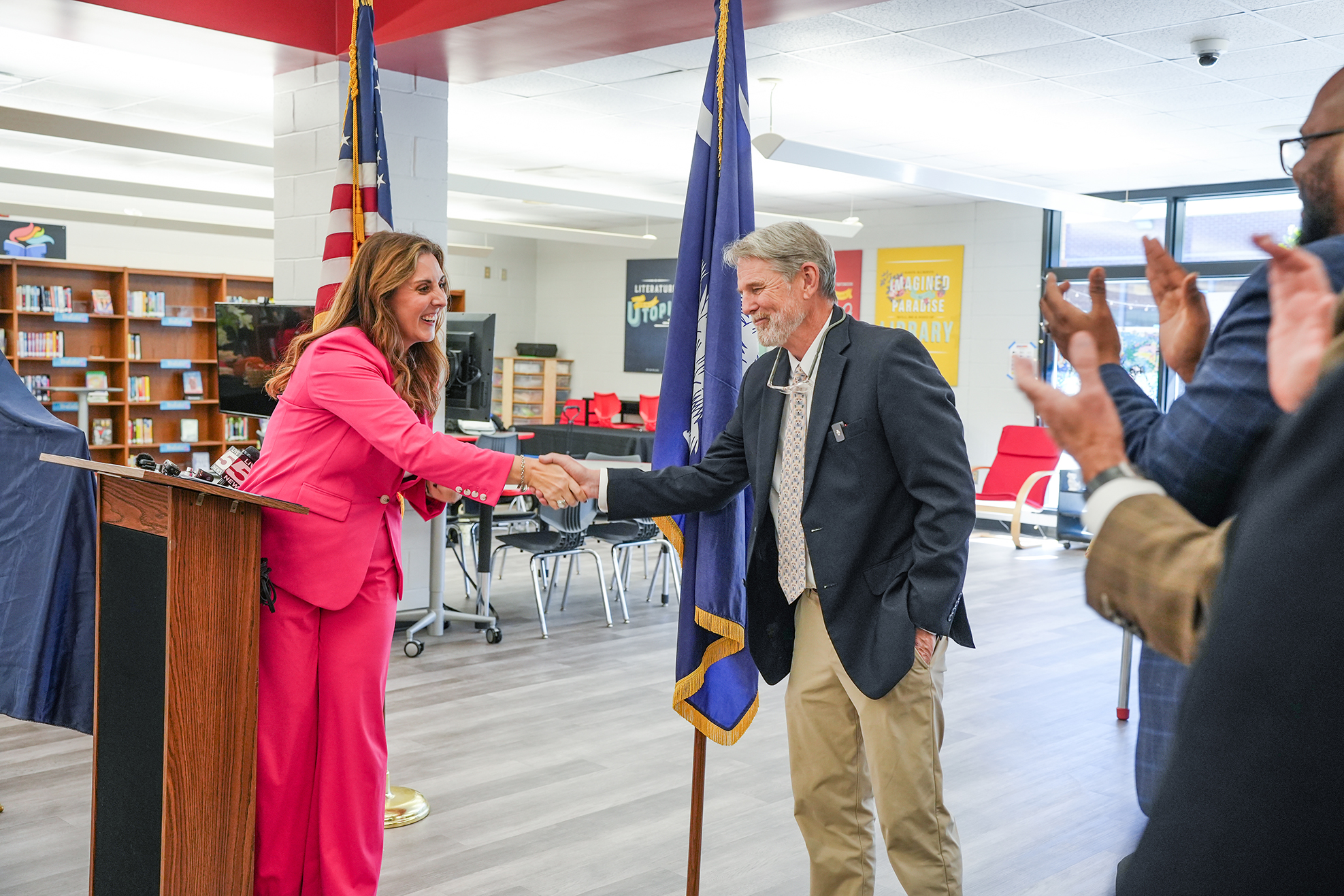 A woman in a pink suit shakes hands with a man in a dark suit. Behind them, three people clap. A podium and flags are present in the room.