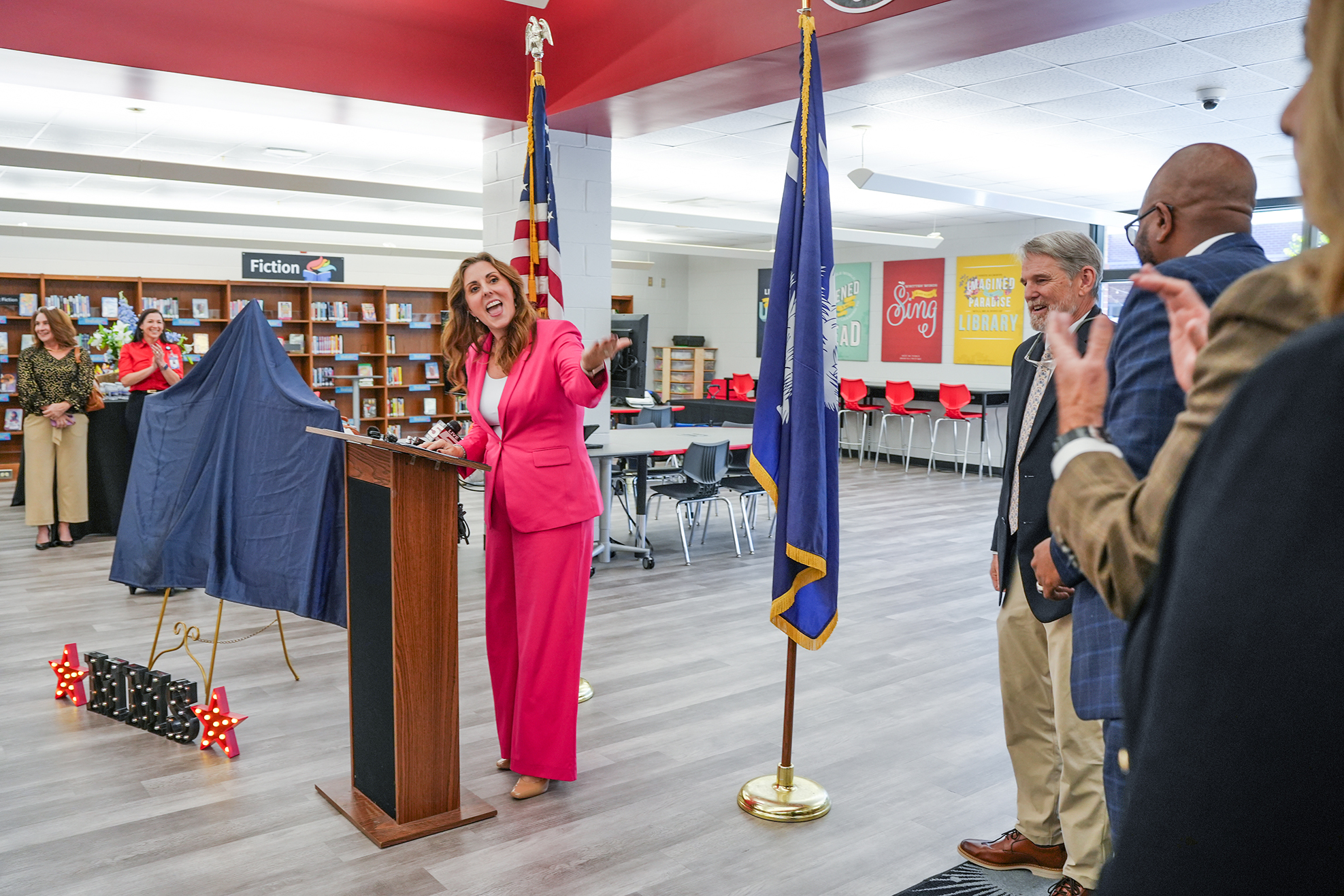 A woman stands at a podium with a flag. A blue cloth covers an object behind her. People listen.
