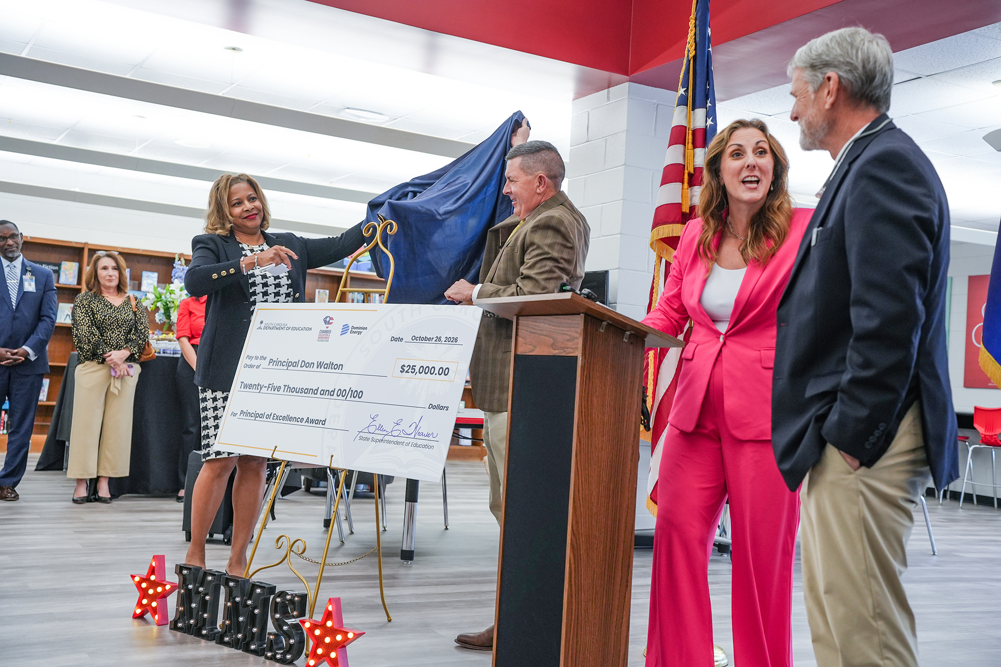 Group of people standing in a room; two women hold a large check and a flag.