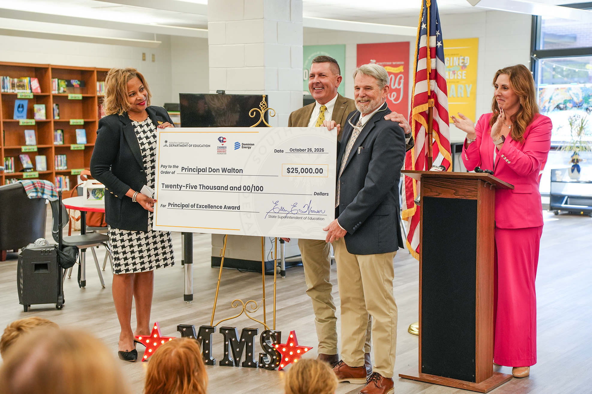 Three adults stand at a podium holding a large check. Behind them is a flag. In the background, there are bookshelves and chairs.