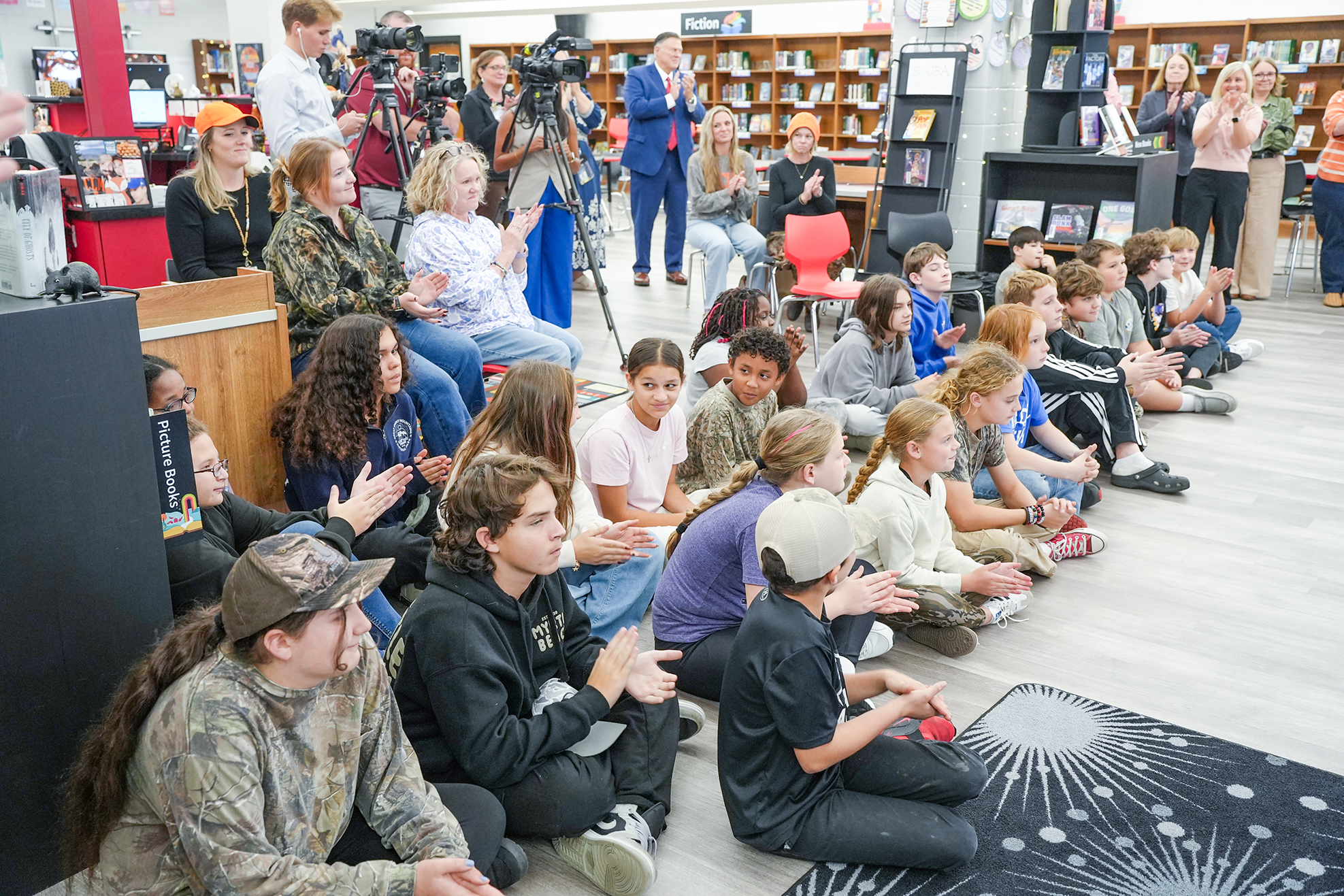 Group of students and teachers sit together in a library, some clap. People stand and hold cameras behind them.