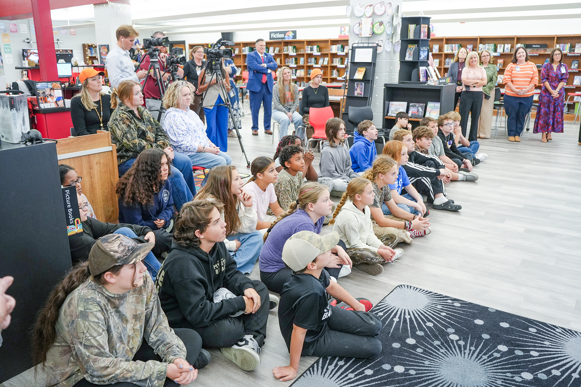 Library with many shelves filled with books. Children sit on floor. Adults stand and sit in back.