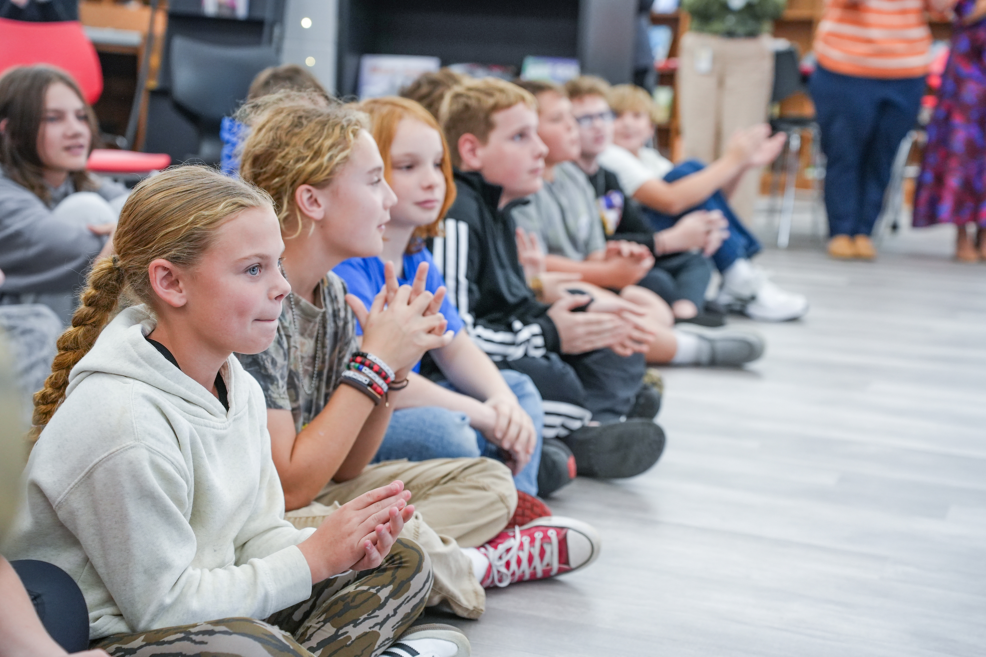 A classroom with children sitting in rows, clapping. A teacher stands in the background.