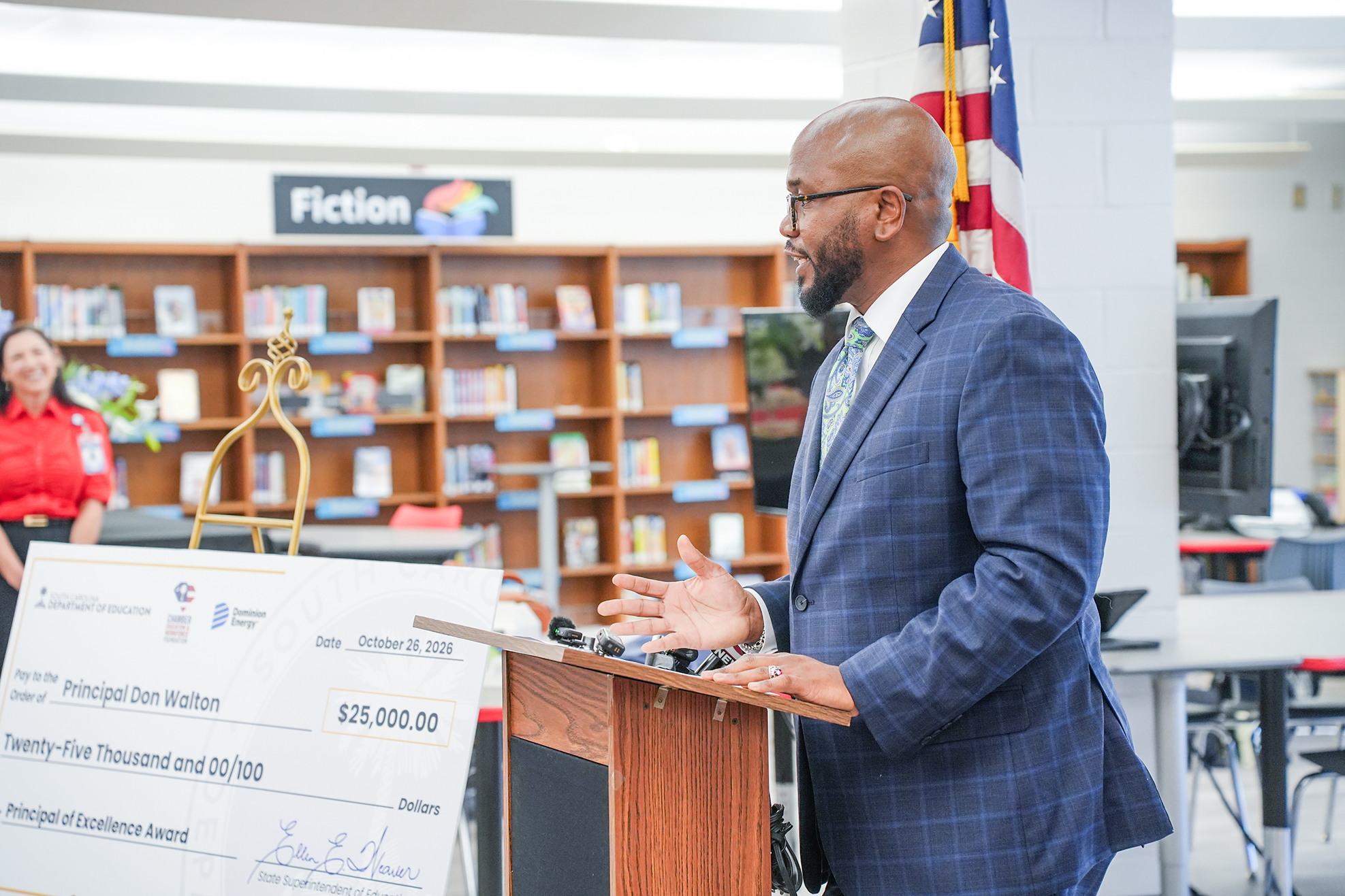 A man wearing a blue suit and glasses speaks at a podium in a classroom.