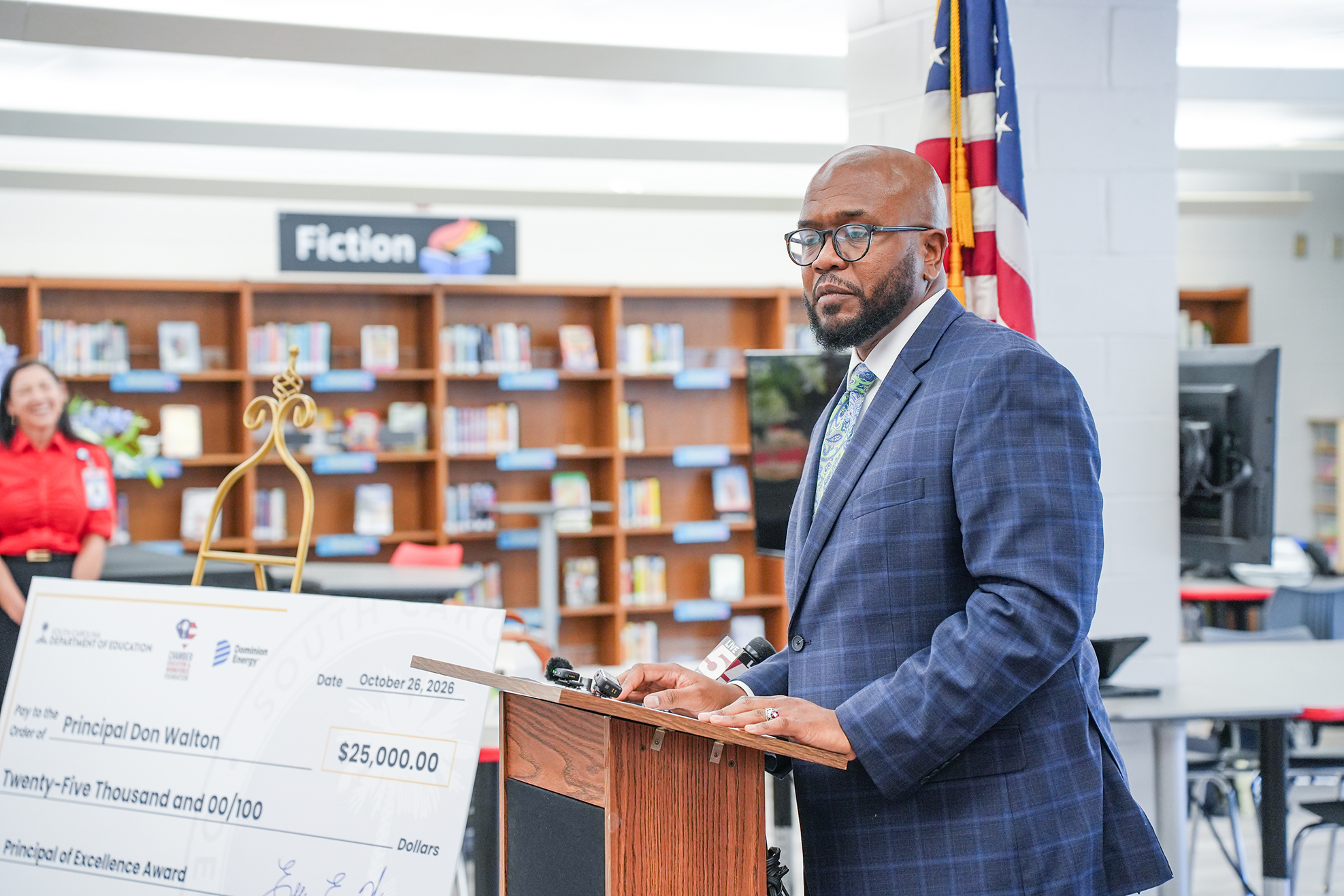A man in a blue suit stands at a podium in a library with bookshelves, holding a microphone.