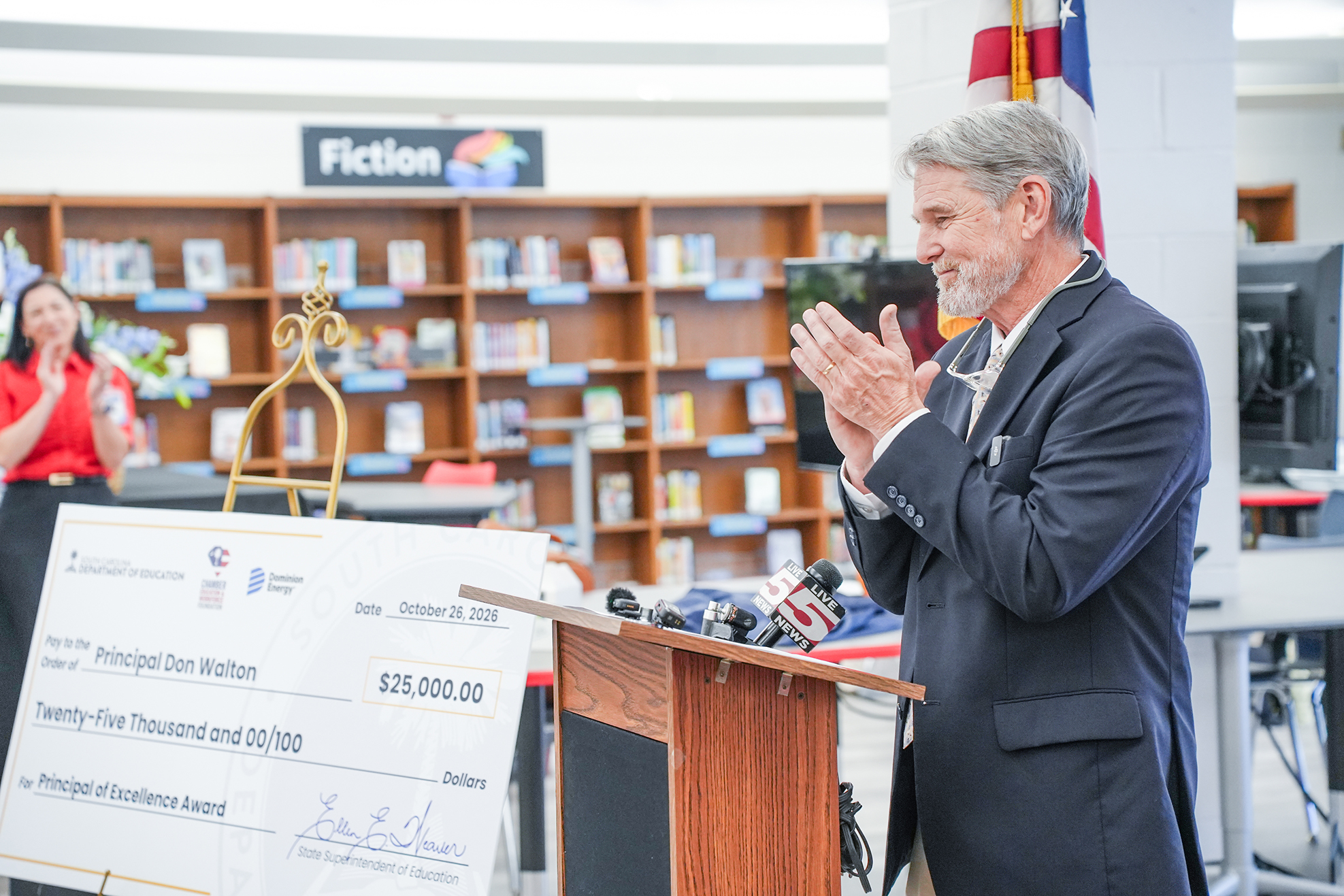 Person with gray hair stands behind a podium in a library with a large check on display.