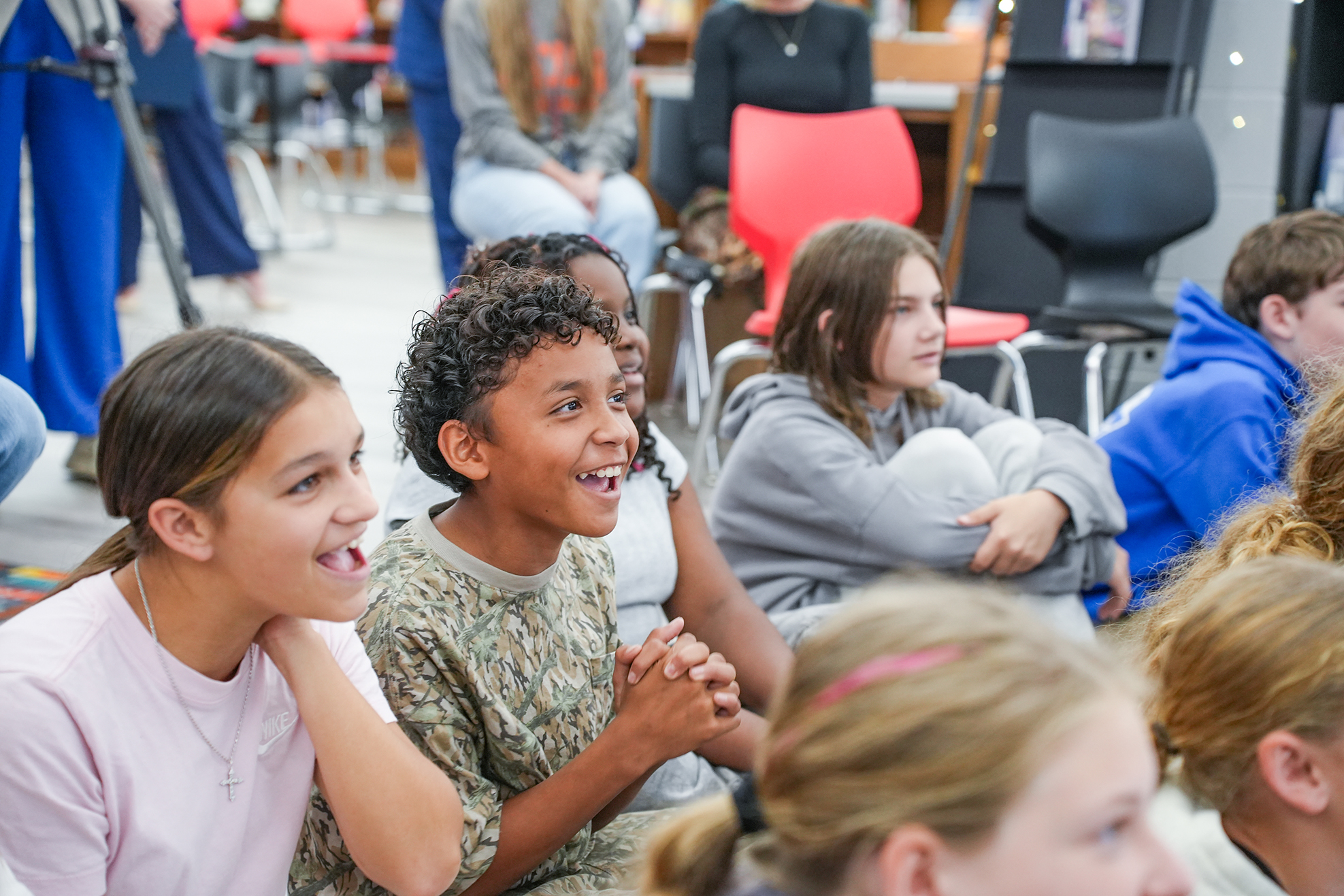 Group of young children in a classroom setting, smiling and sitting on floor, some with arms crossed, others clapping.
