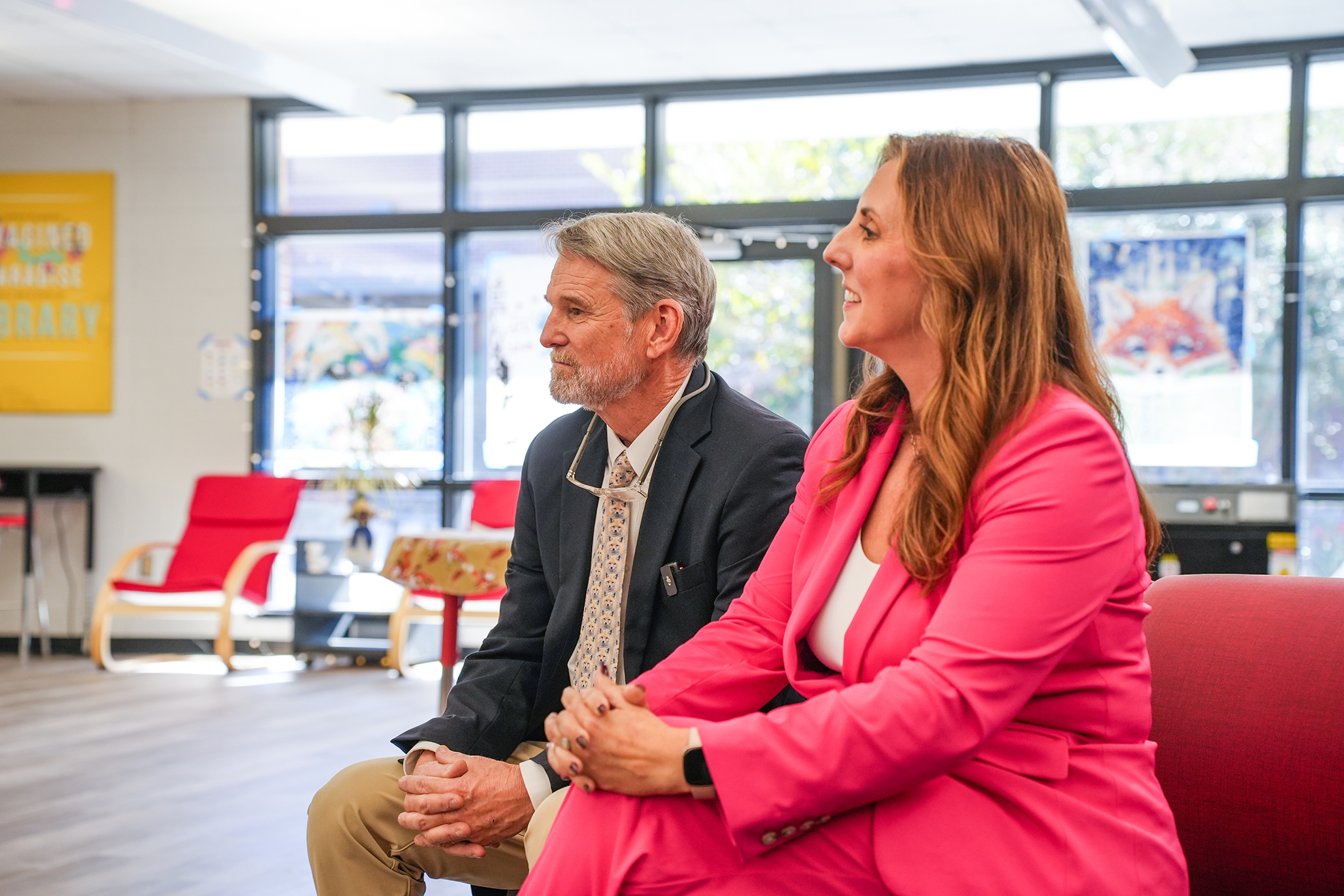 Two adults sitting on chairs, smiling and talking; woman wears pink blazer, man wears glasses and dark suit; yellow wall with white text behind them