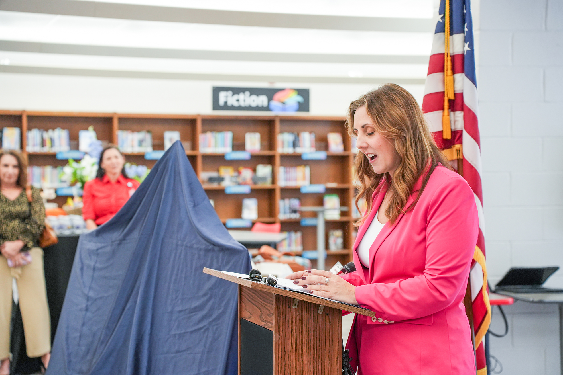 Woman in pink blazer reading aloud at podium with American flag behind her, library in background.