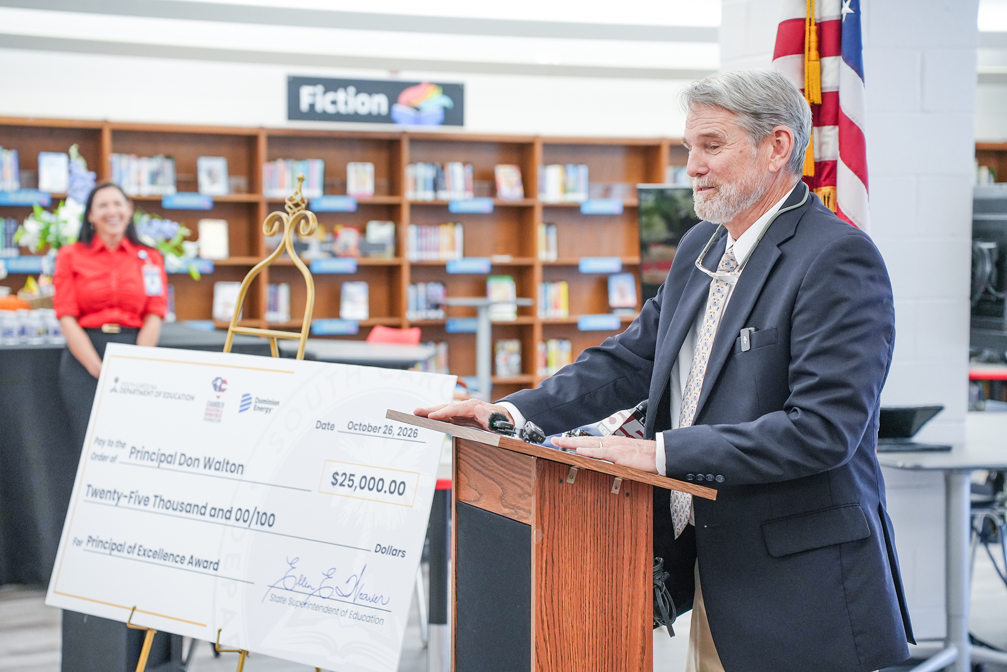 A man stands at a podium reading a large check. Behind him, a woman stands near a shelf full of books.
