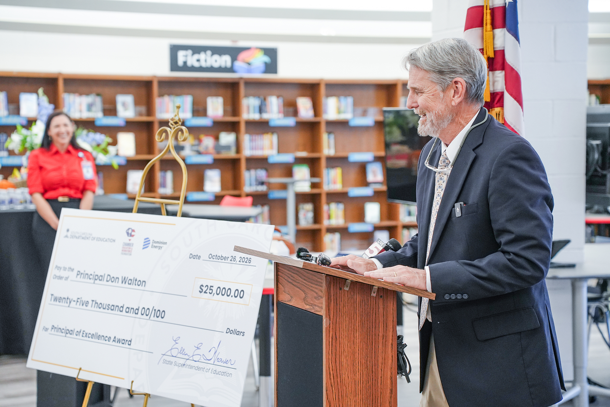 Man in suit at a podium with a large check in front of him. Woman in red shirt stands behind him.